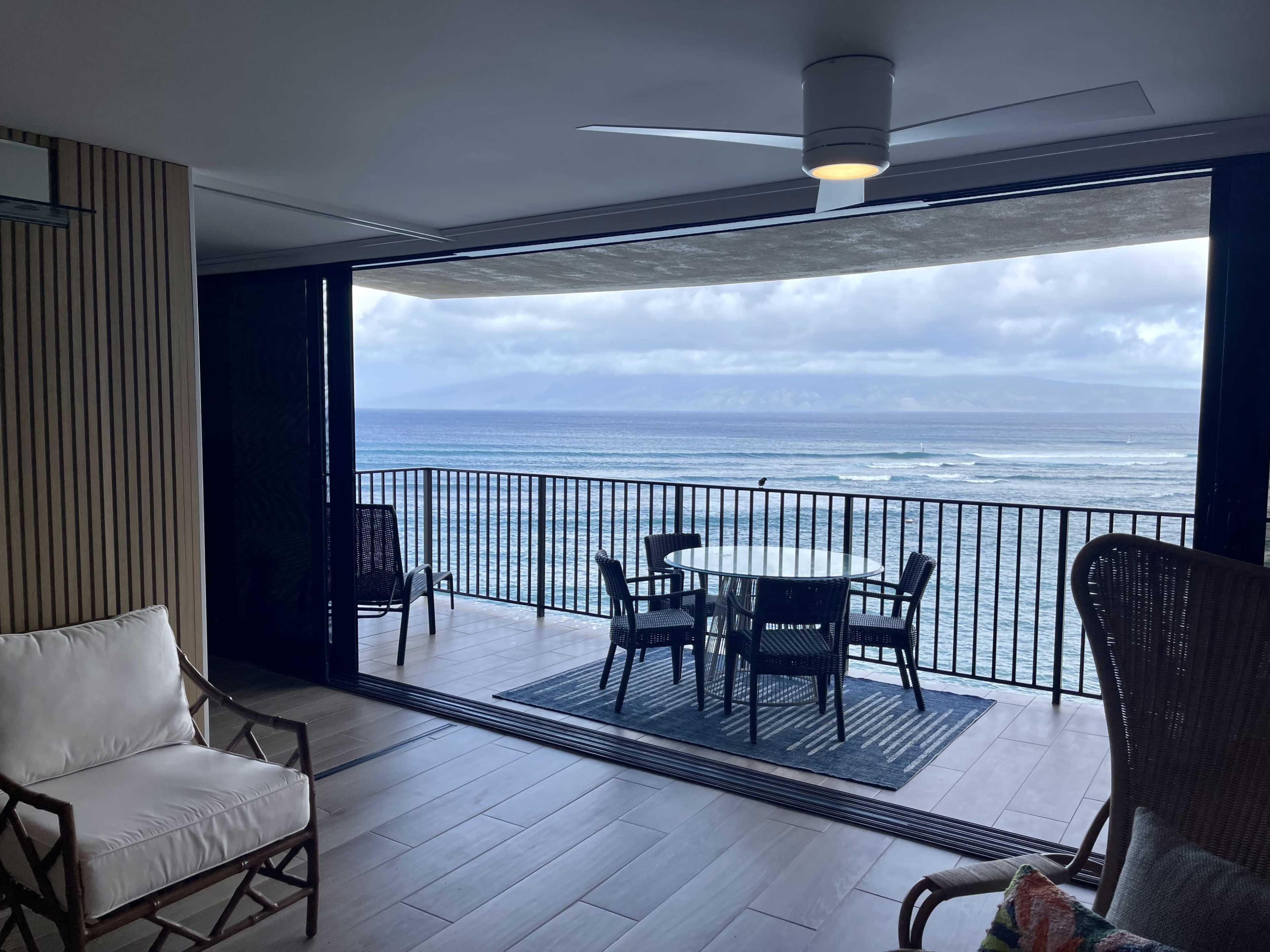A seating area with chairs and a table overlooks a beach and ocean through a large open sliding door.