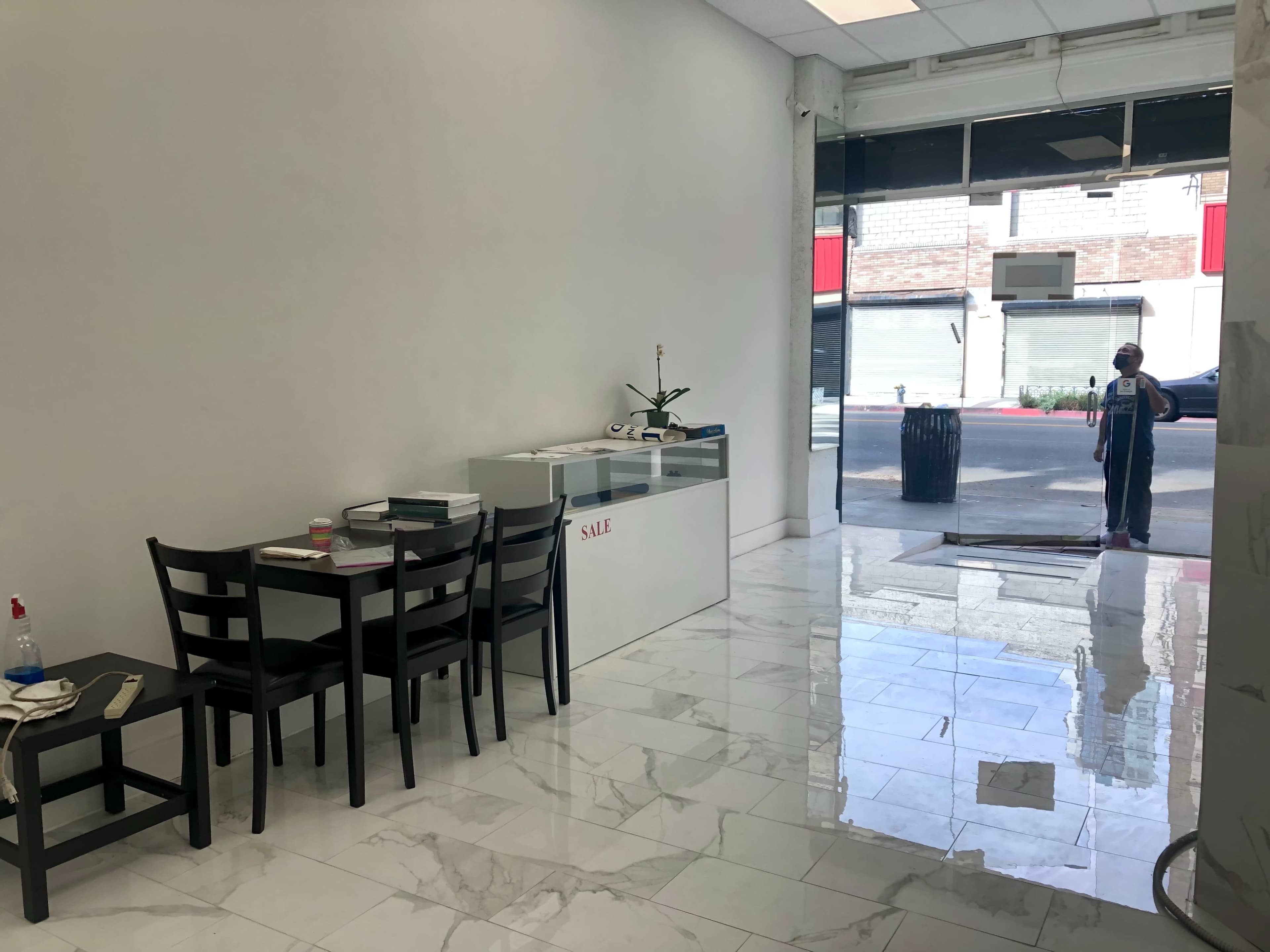 A clean, empty retail space features a display counter and a few black chairs near the entrance, with a person standing outside.