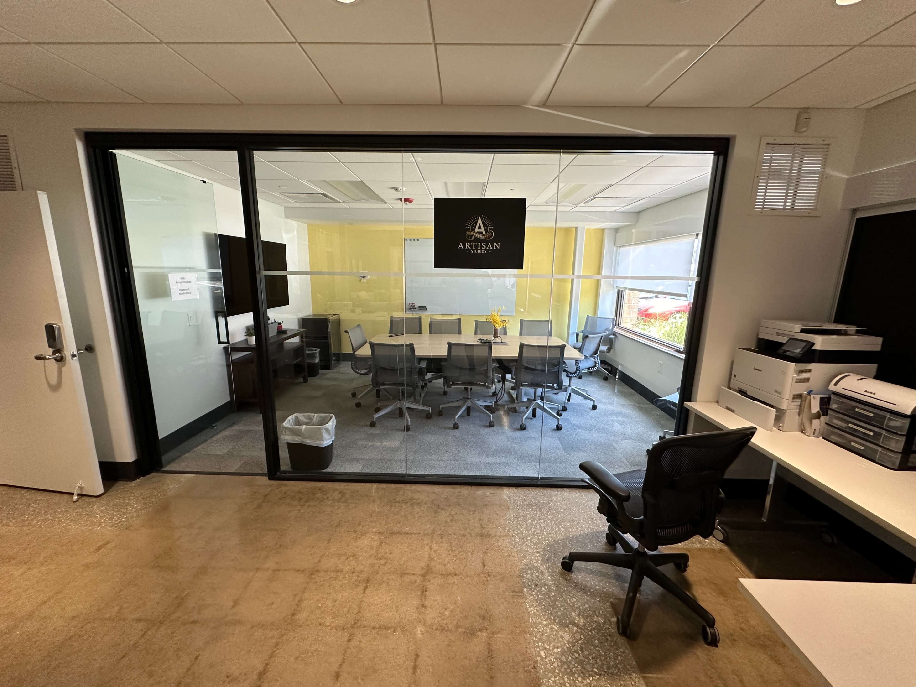 A modern conference room with glass walls, featuring a long table surrounded by ergonomic chairs and a large television mounted on the wall.