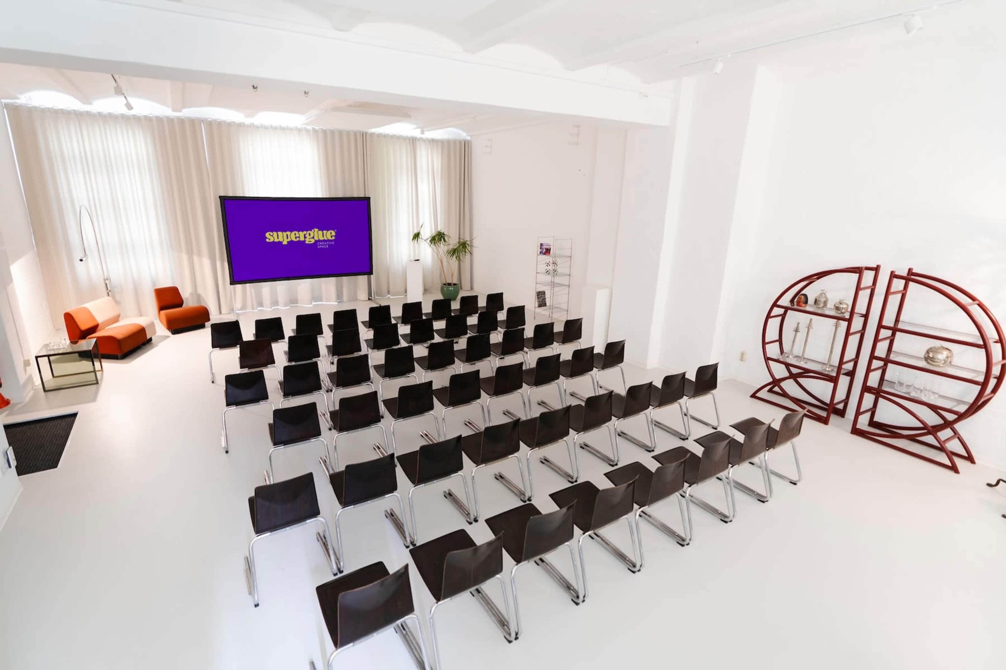 The image shows a modern meeting room with rows of black chairs facing a large screen displaying the word "superglue," accompanied by orange seating and a decorative bookshelf.