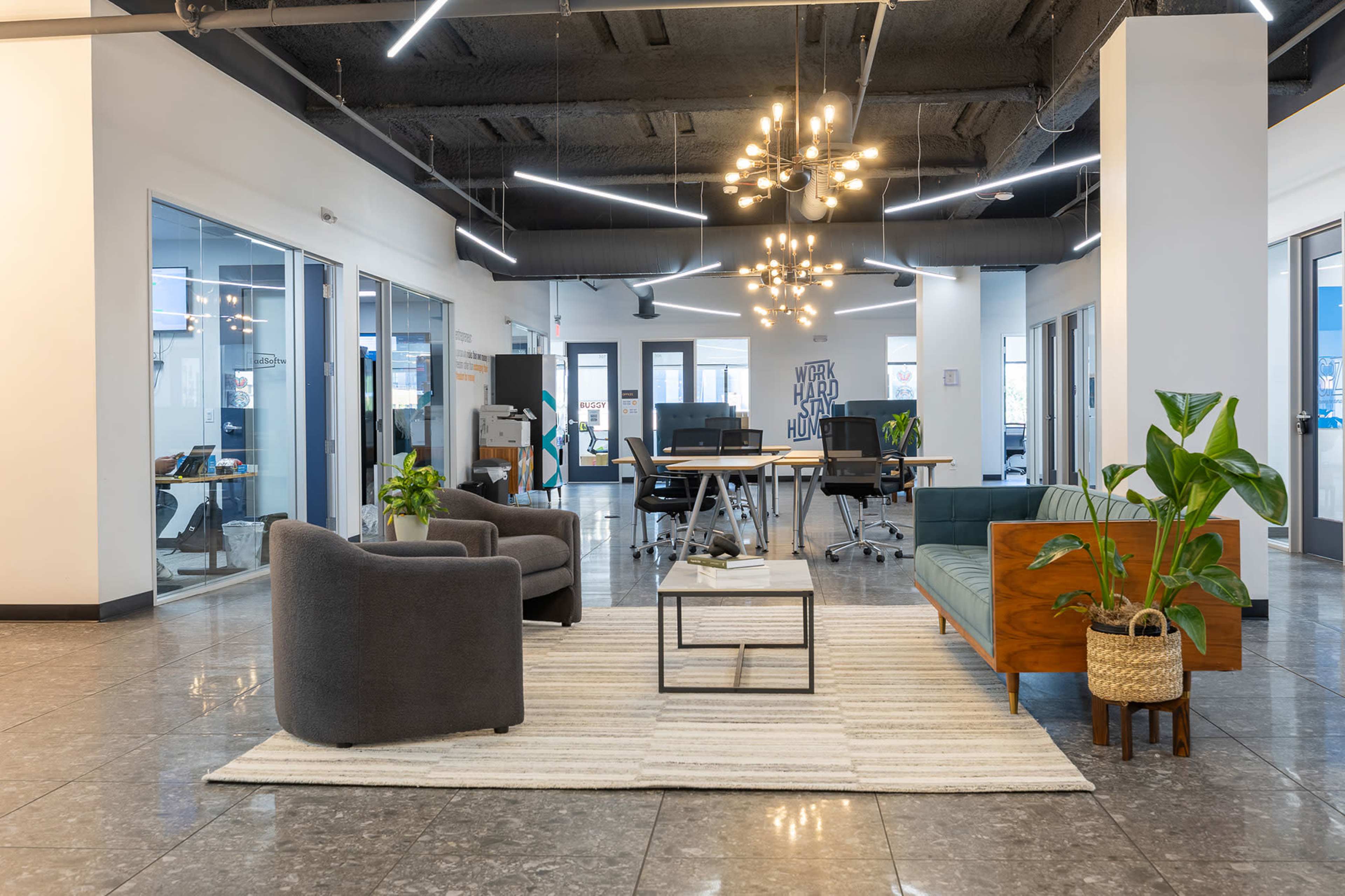 The image depicts a modern office space featuring a seating area with a couch, armchairs, and a coffee table, surrounded by meeting rooms and workspaces.