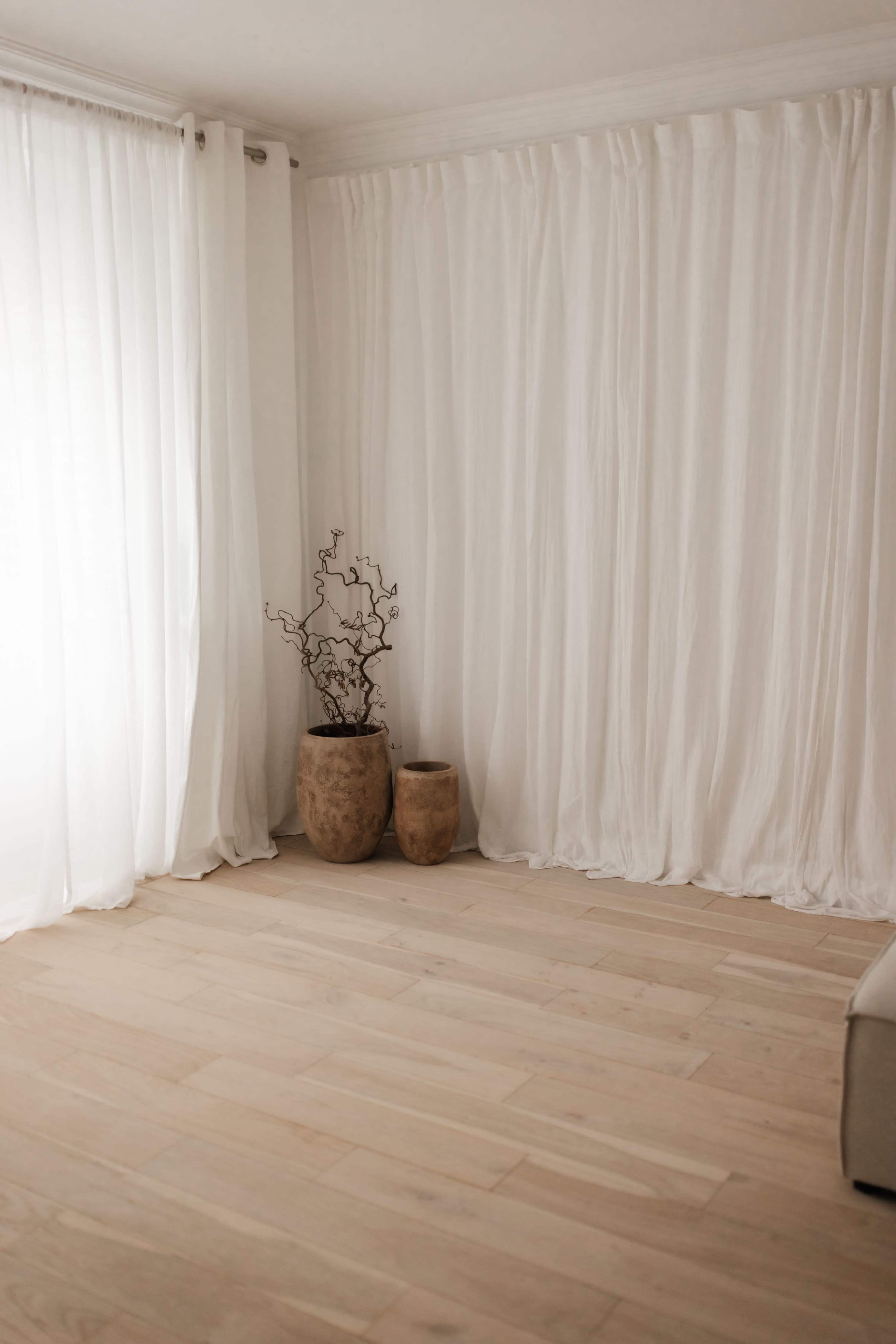 The image shows a bright room with white curtains, wooden flooring, and two decorative vases containing branches.