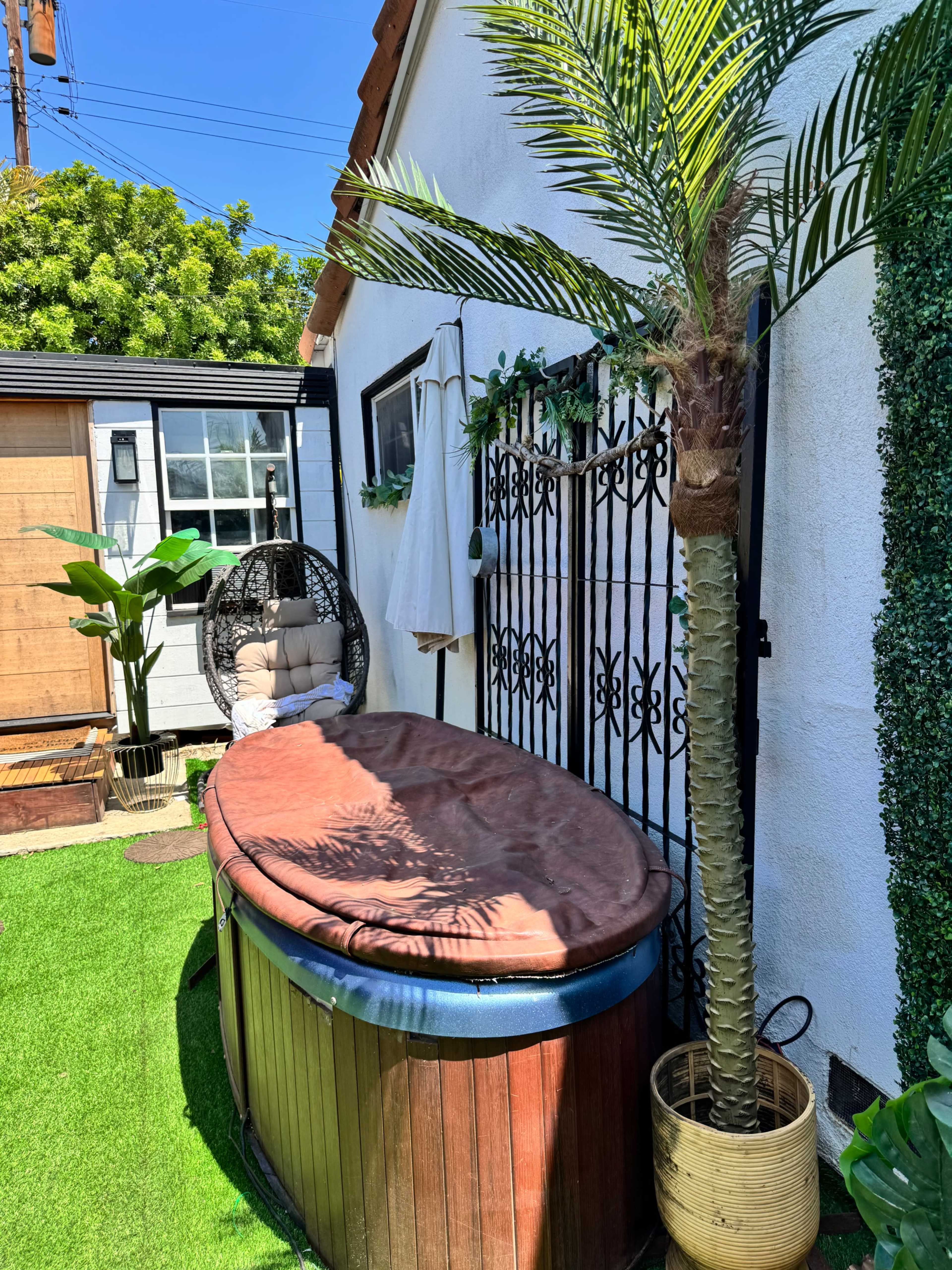 The image shows a wooden hot tub with a brown cover placed next to a small palm tree and a hanging chair in a well-maintained outdoor space.