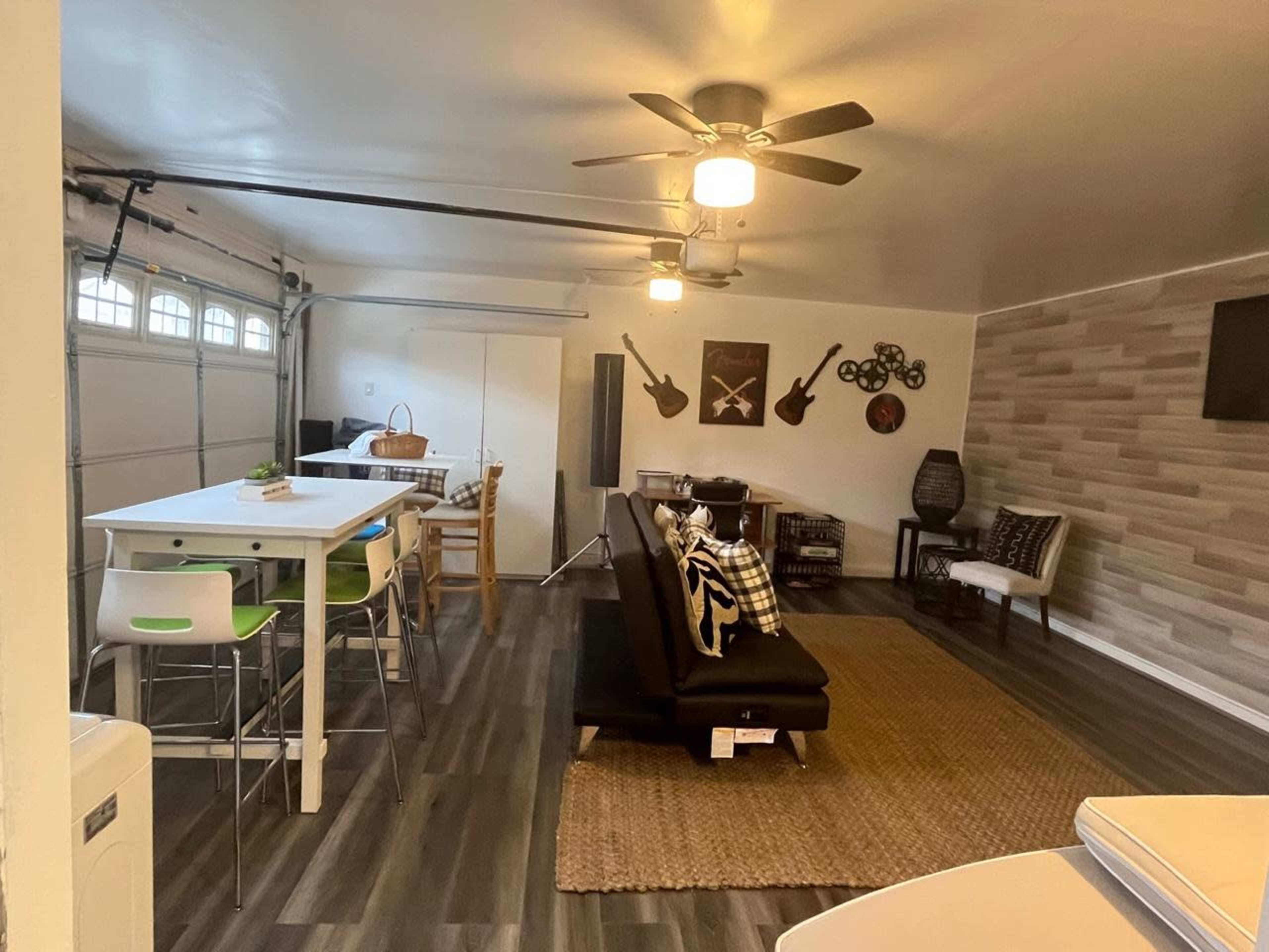 A modern, open-concept room featuring a dining area with tables and chairs, a living space with a loveseat, and a garage door, all illuminated by ceiling lights.