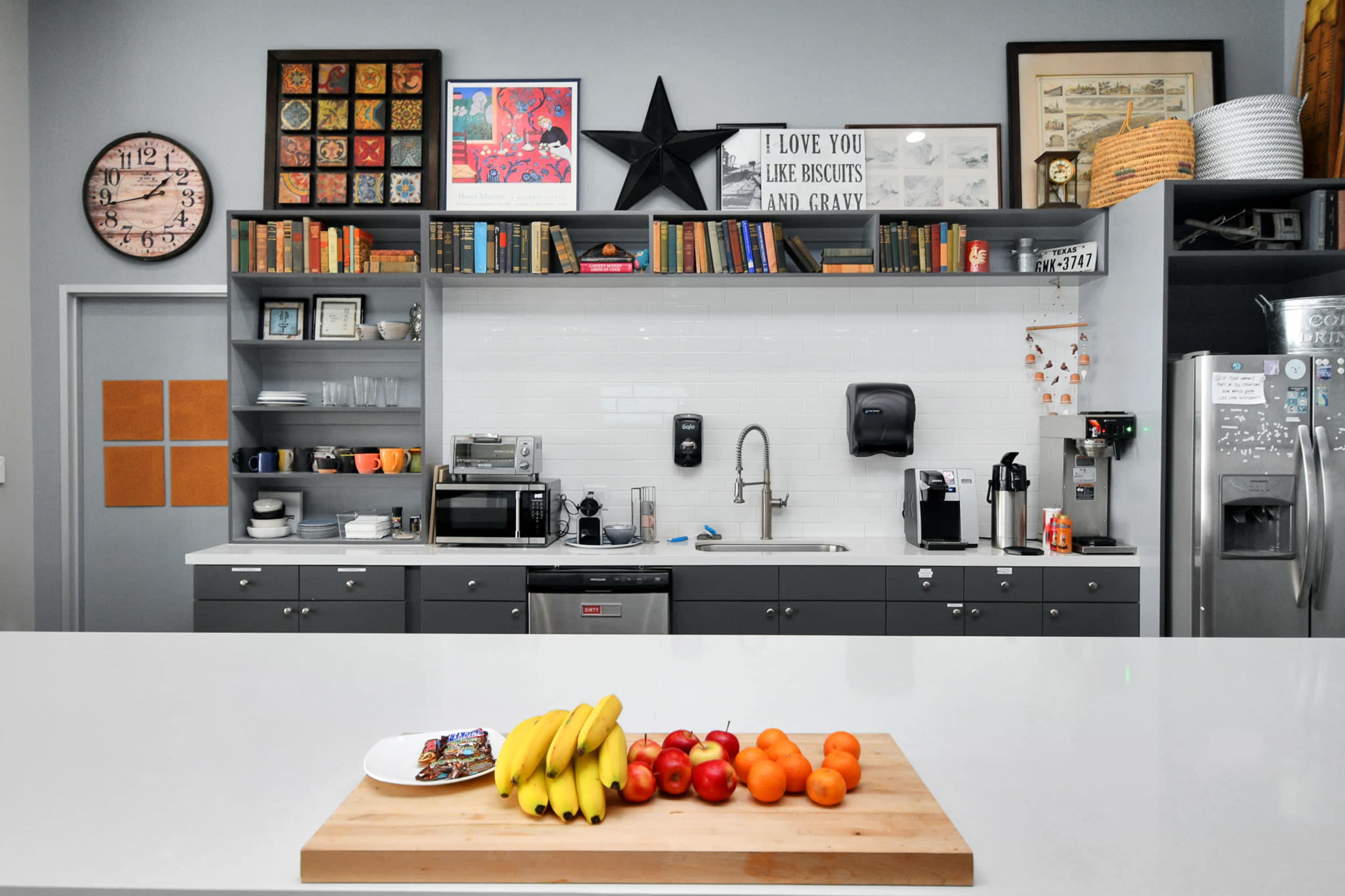 The image shows a modern kitchen featuring a central island with a cutting board holding bananas and apples, and a wall decorated with various framed artwork and shelves filled with books and kitchenware.
