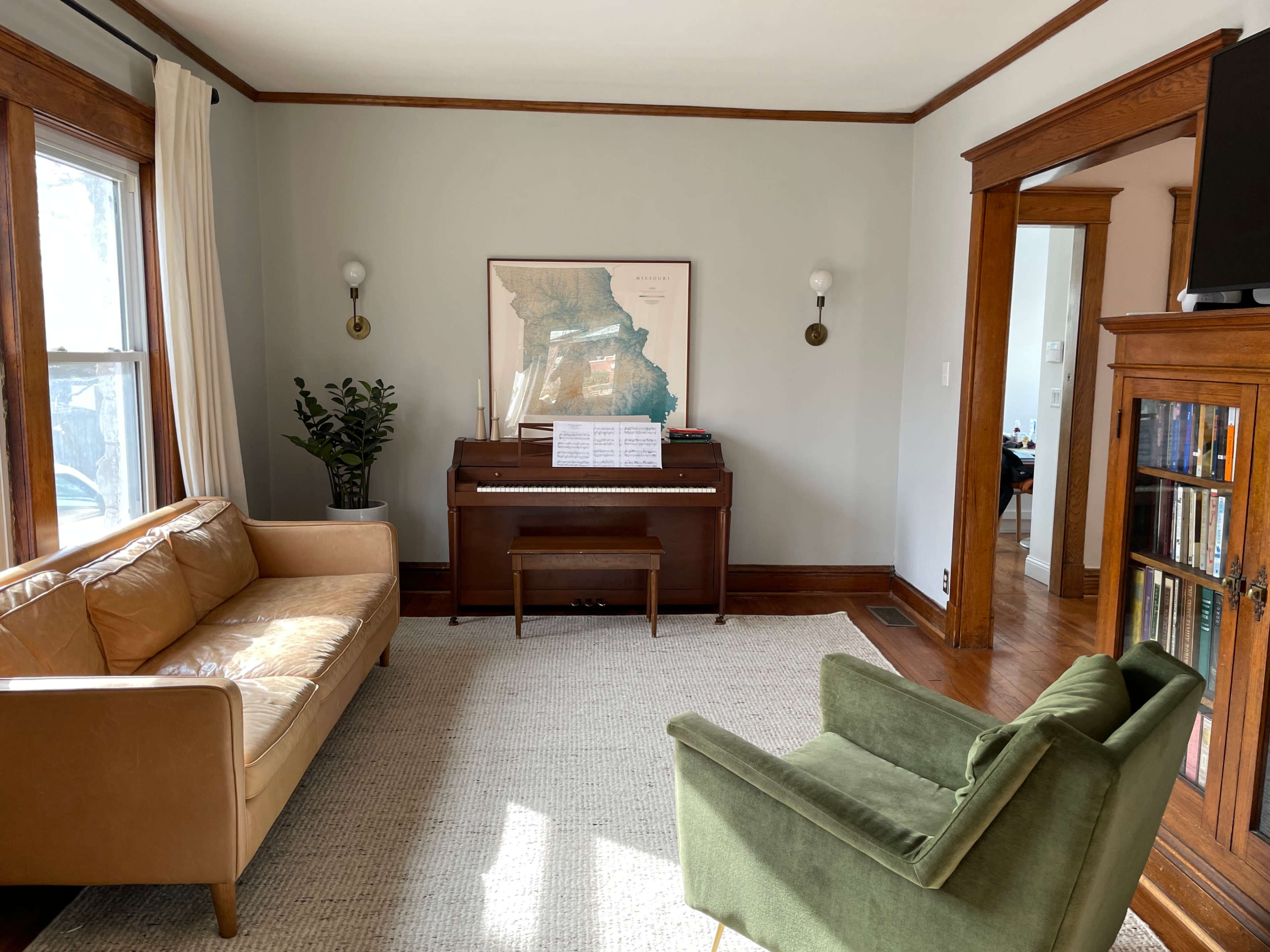 The image shows a cozy living room with a brown leather sofa, a green armchair, a piano with sheet music, and a large map framed on the wall.