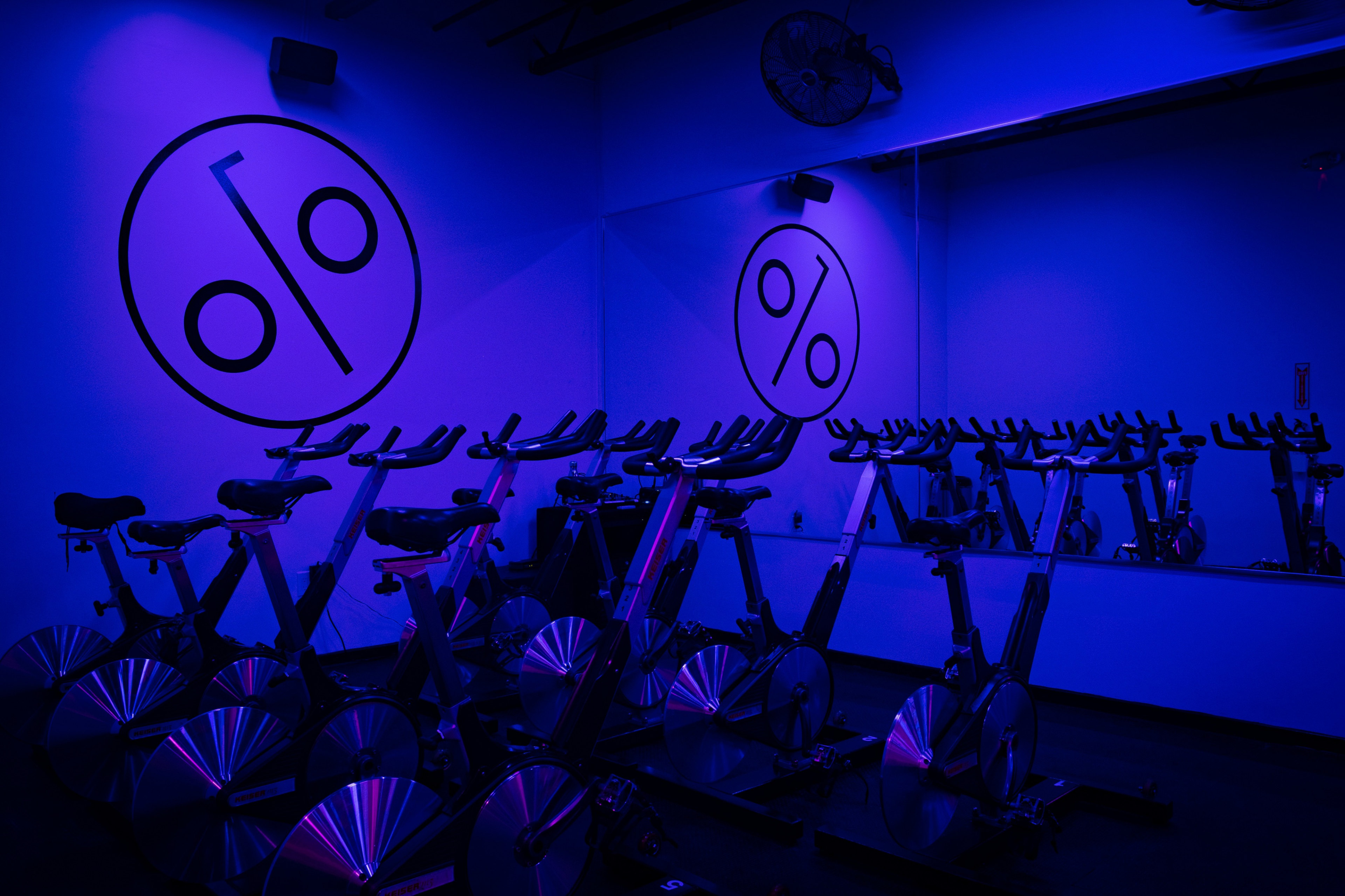 A dimly lit indoor cycling studio features multiple stationary bikes against a backdrop of large, circular logos on the walls.