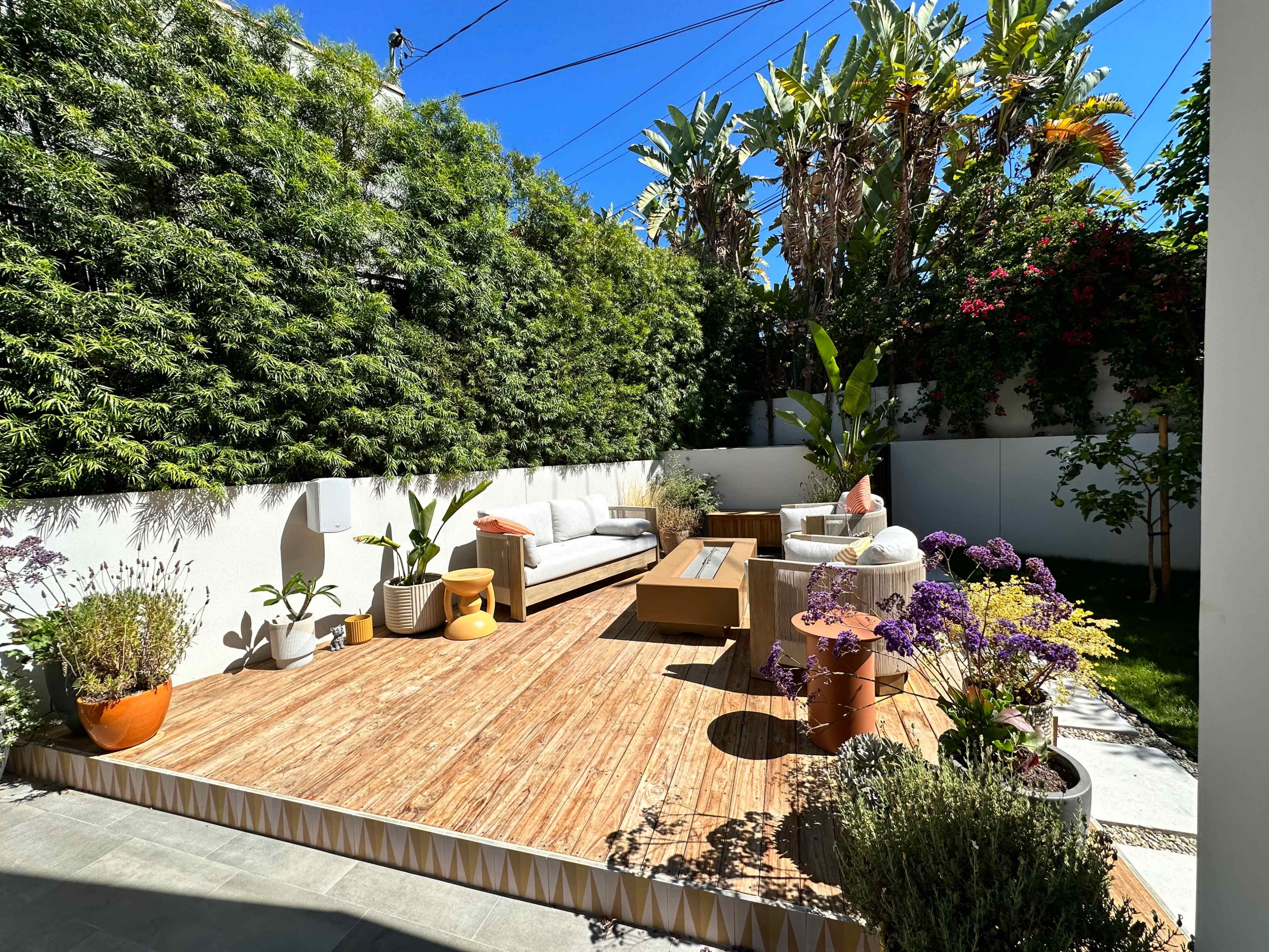 A modern outdoor patio features wooden decking, a sofa, coffee table, and various potted plants amidst lush greenery.