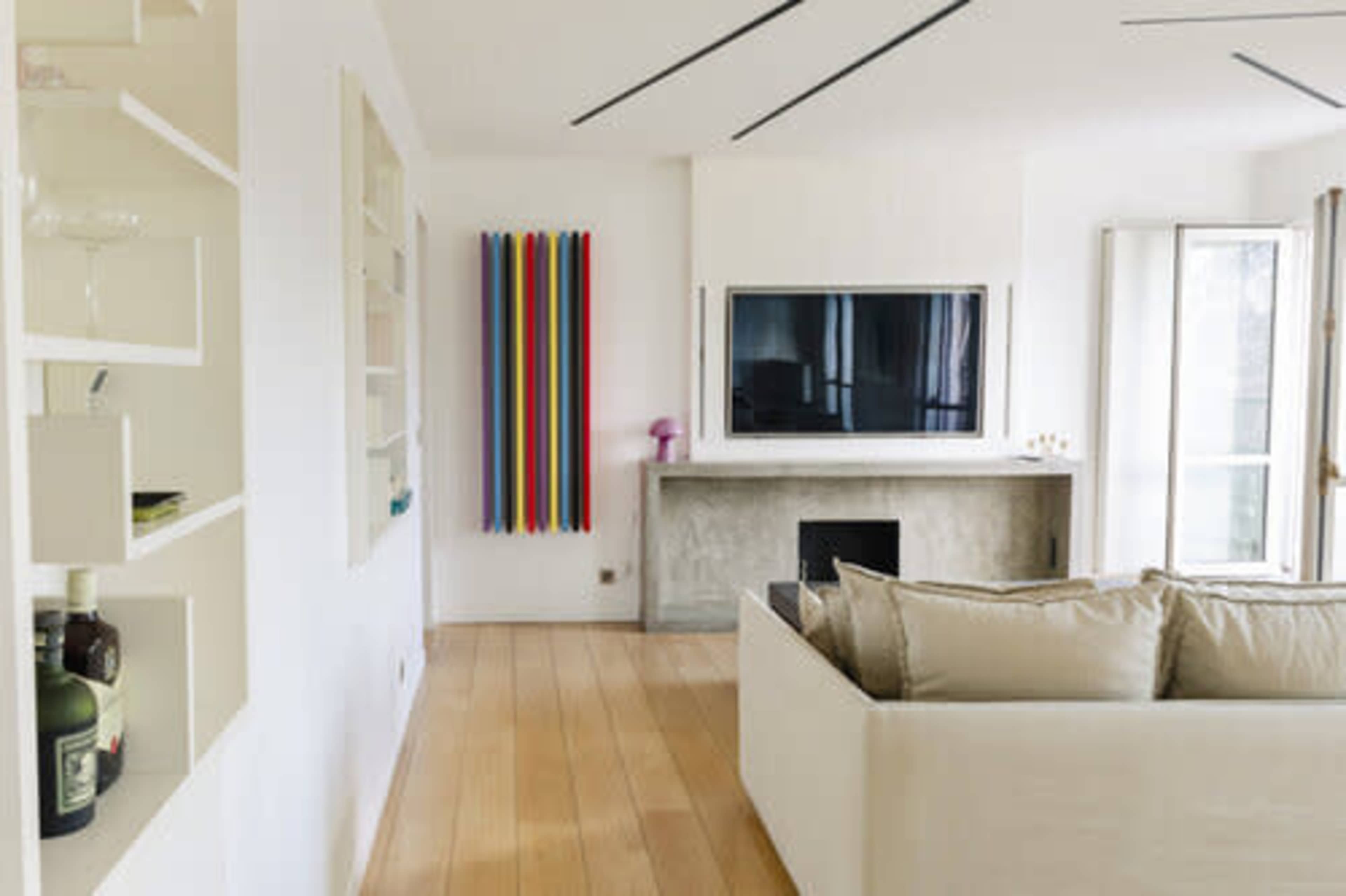 The image shows a modern living room with a white sofa, a television mounted on a wall, and colorful decorative elements arranged vertically.