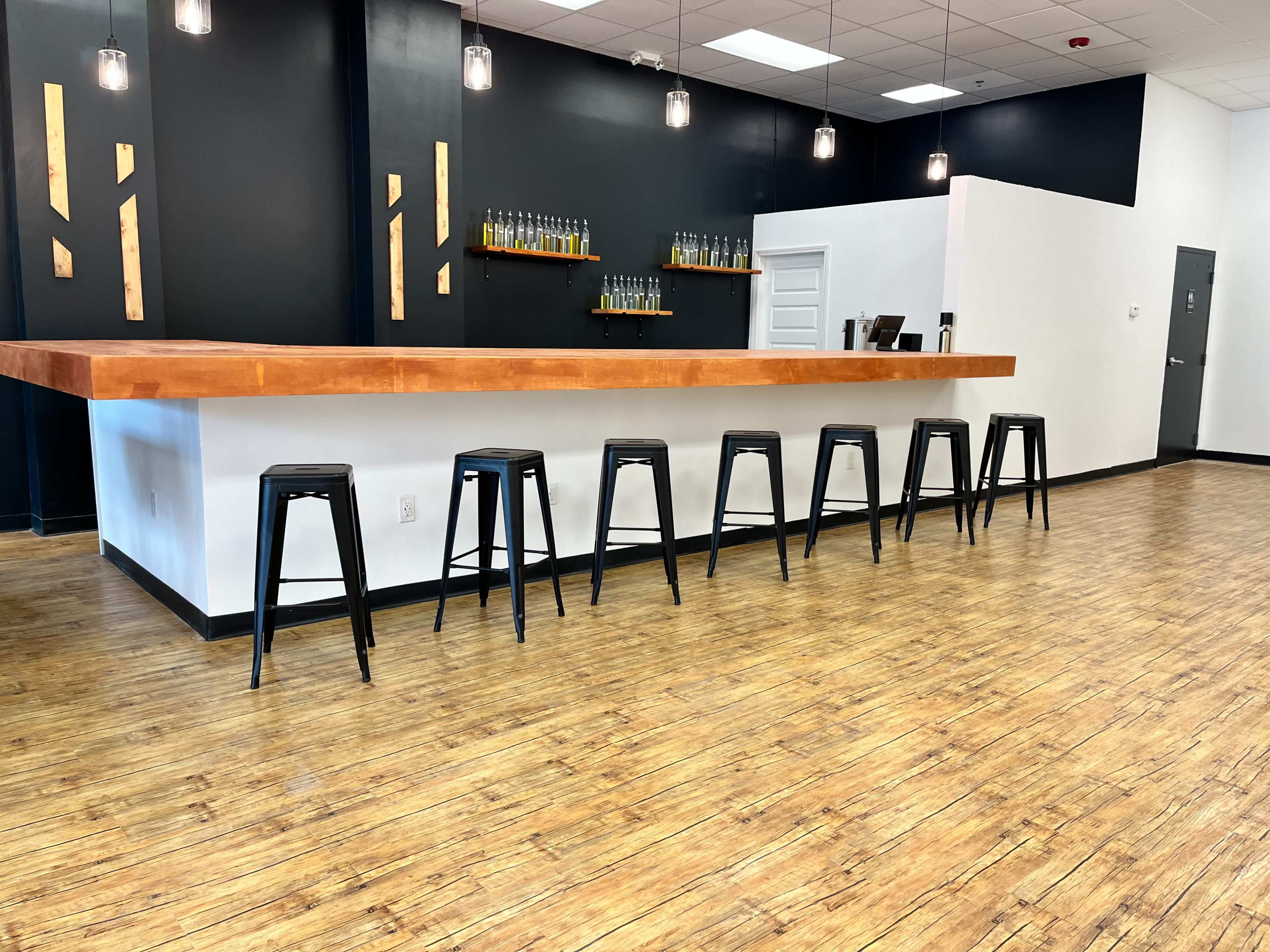 The image shows a minimalist bar area with a wooden countertop, several black stools, and a wall shelf displaying bottles, against a dark background.