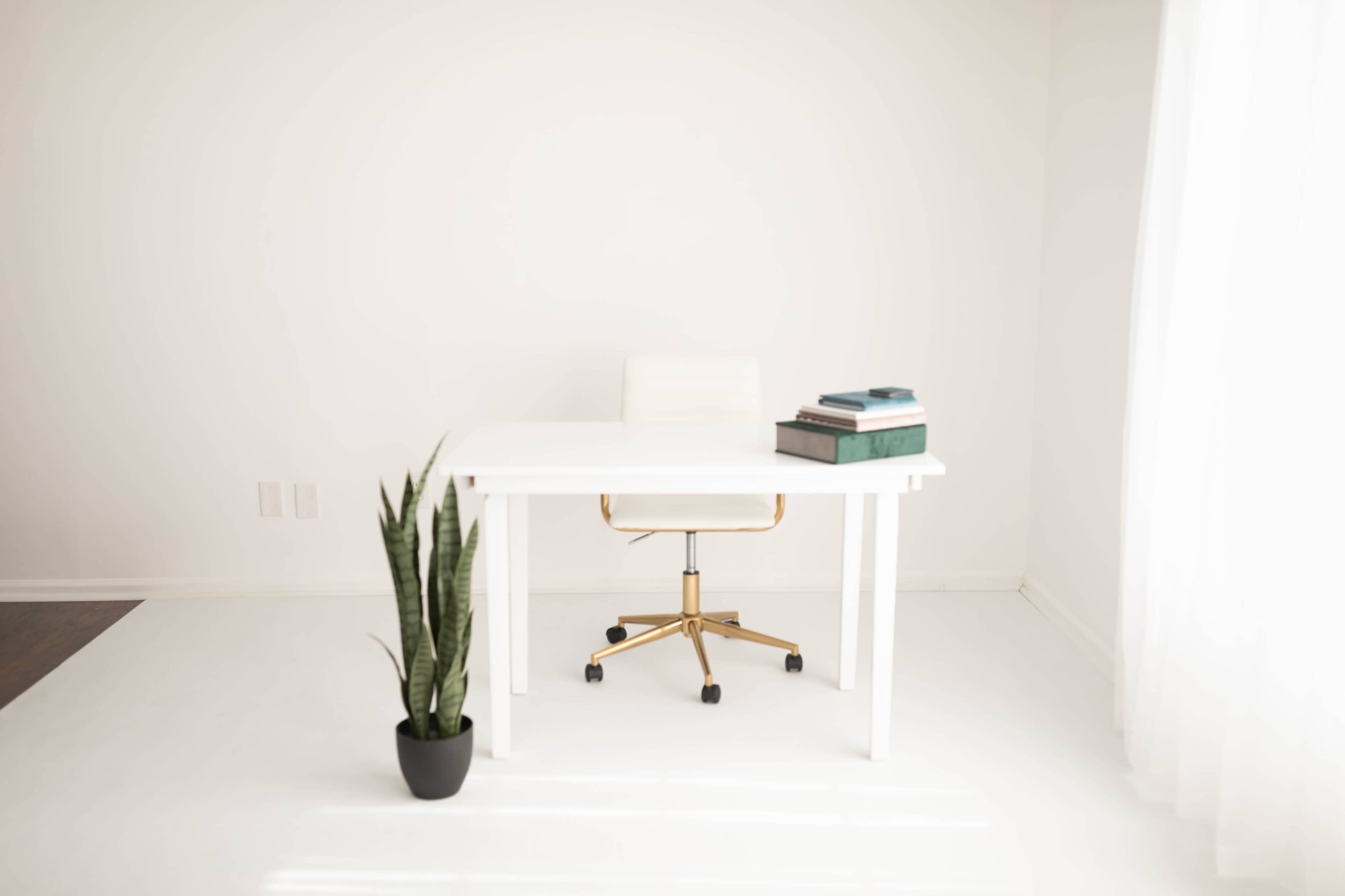 A modern, minimalistic office space features a white desk with a gold rolling chair, stacked books, and a potted plant in a corner.