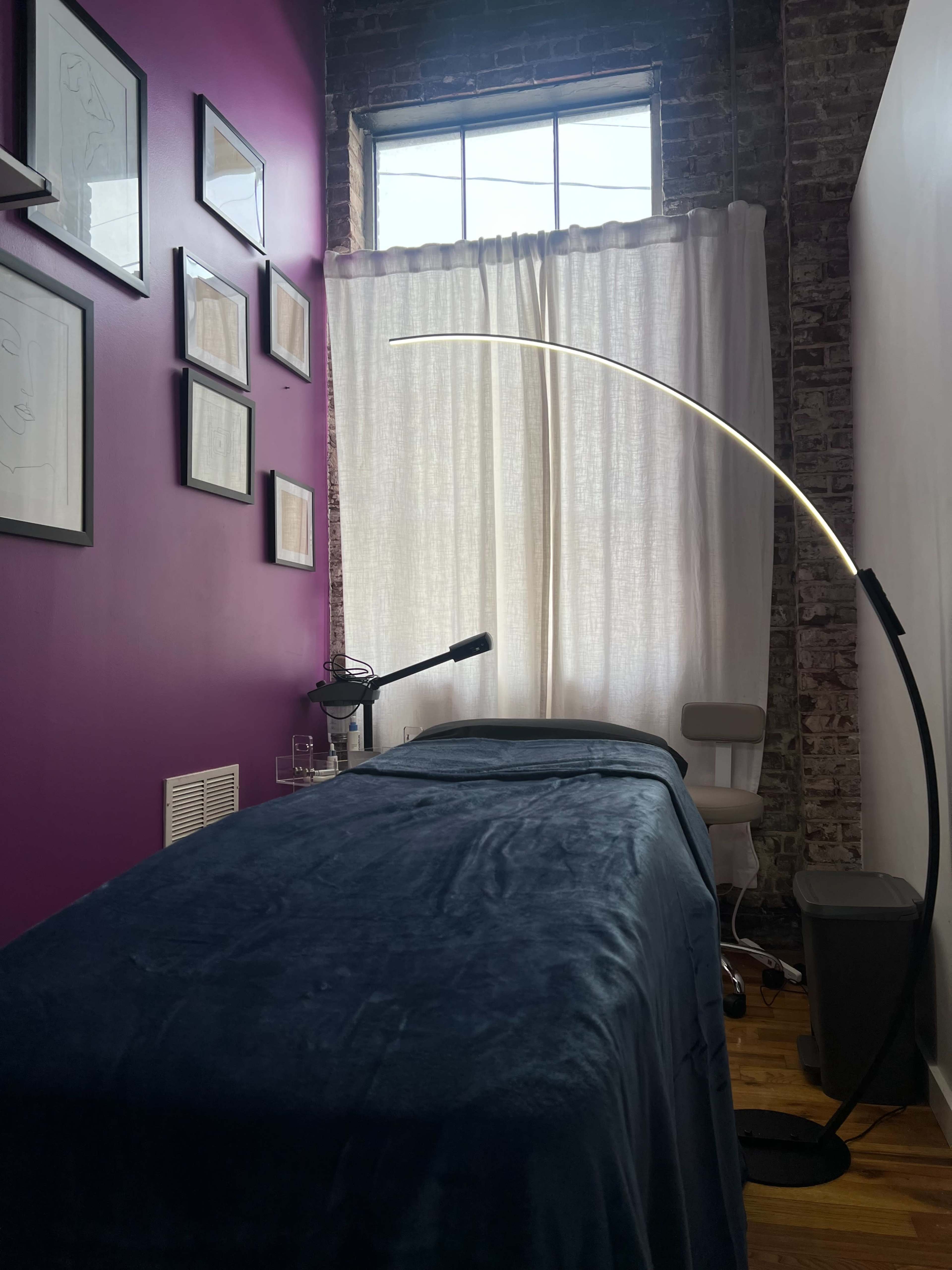 A room with a purple wall featuring framed artwork, a massage table covered with a blue blanket, and a tall lamp beside a chair near a window with sheer curtains.
