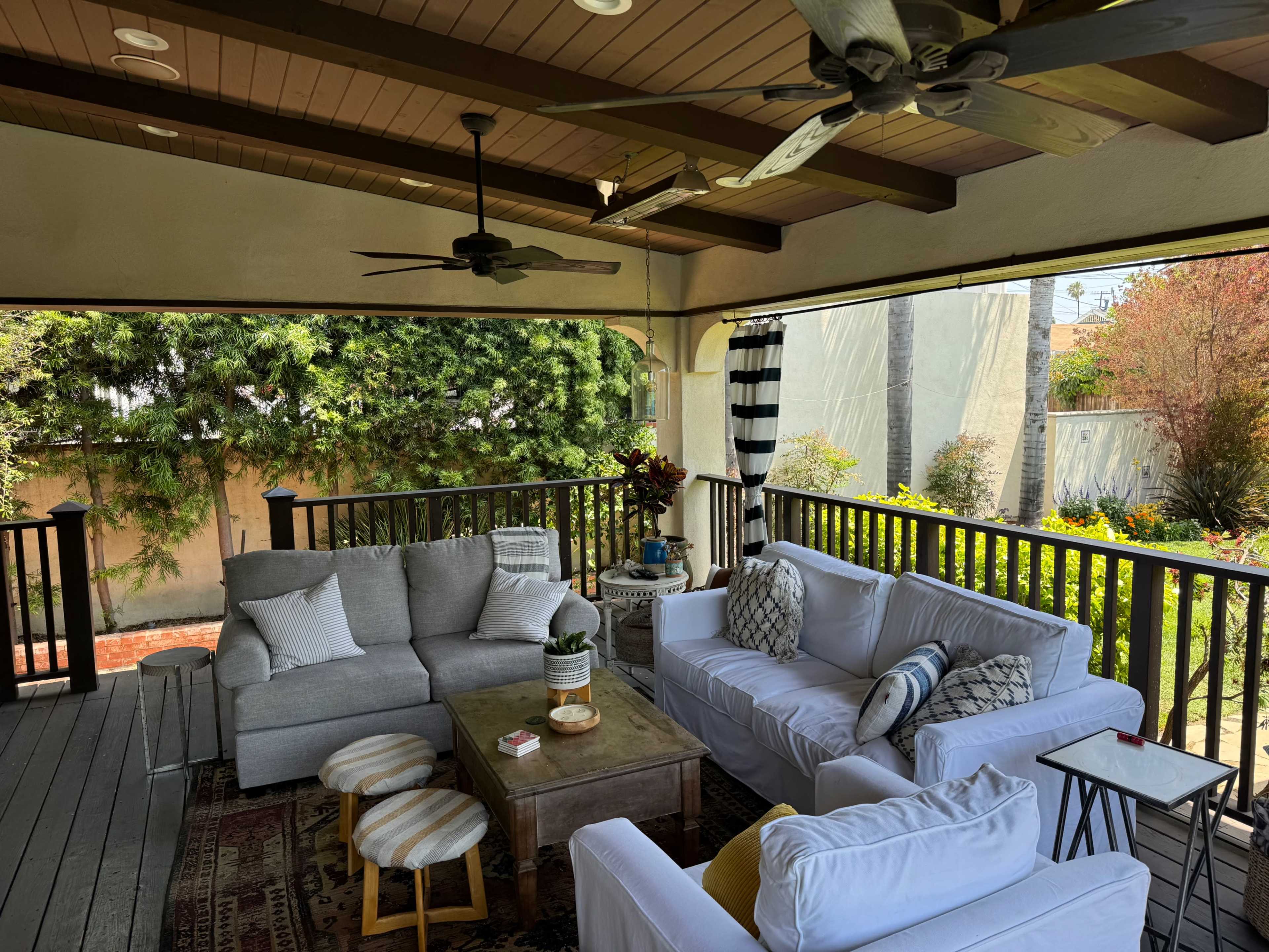 A cozy outdoor seating area features a gray sectional sofa, white chairs, a coffee table, and lush greenery in the background.