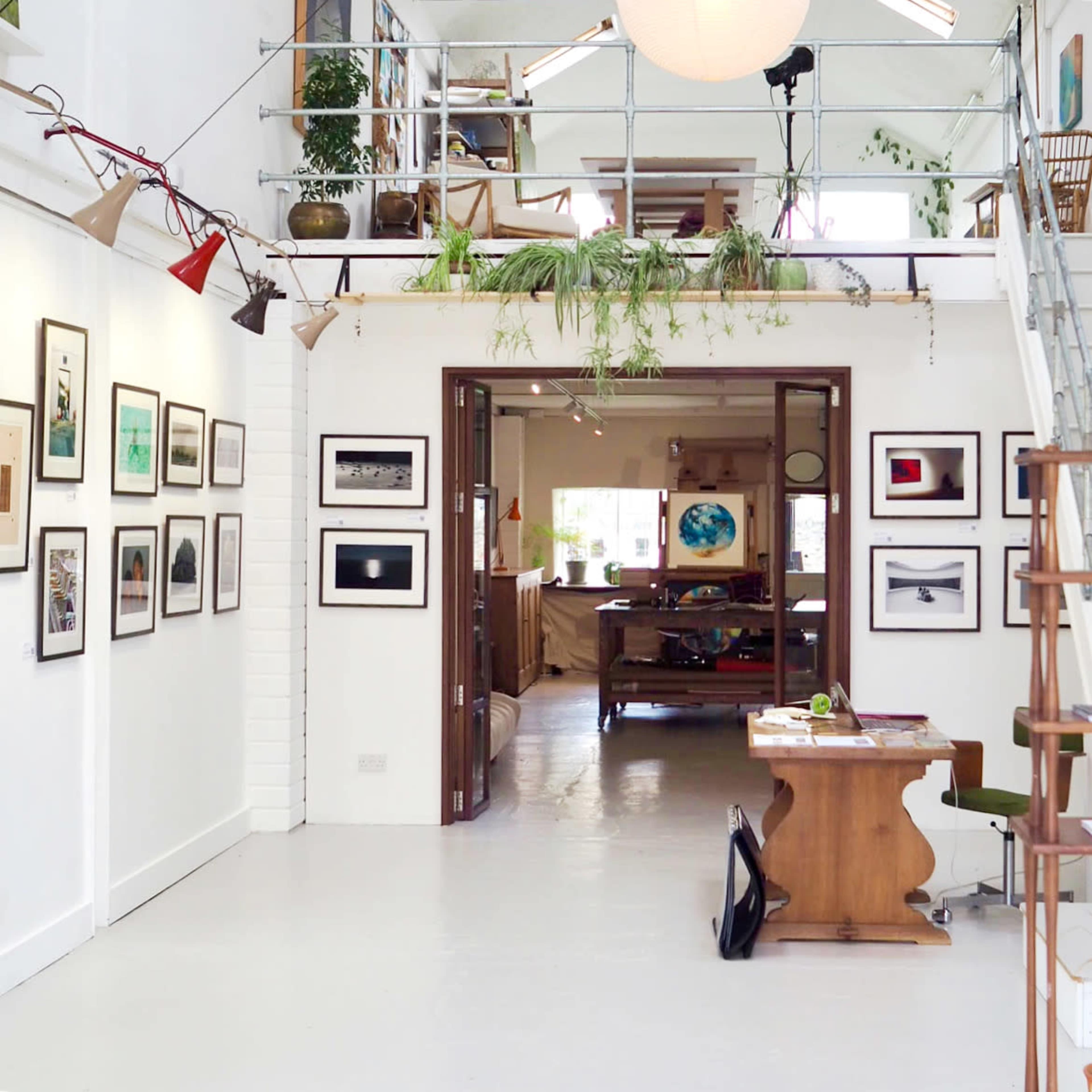 The image shows an art studio with white walls adorned with framed photographs, leading into a spacious area featuring wooden furniture and plants.