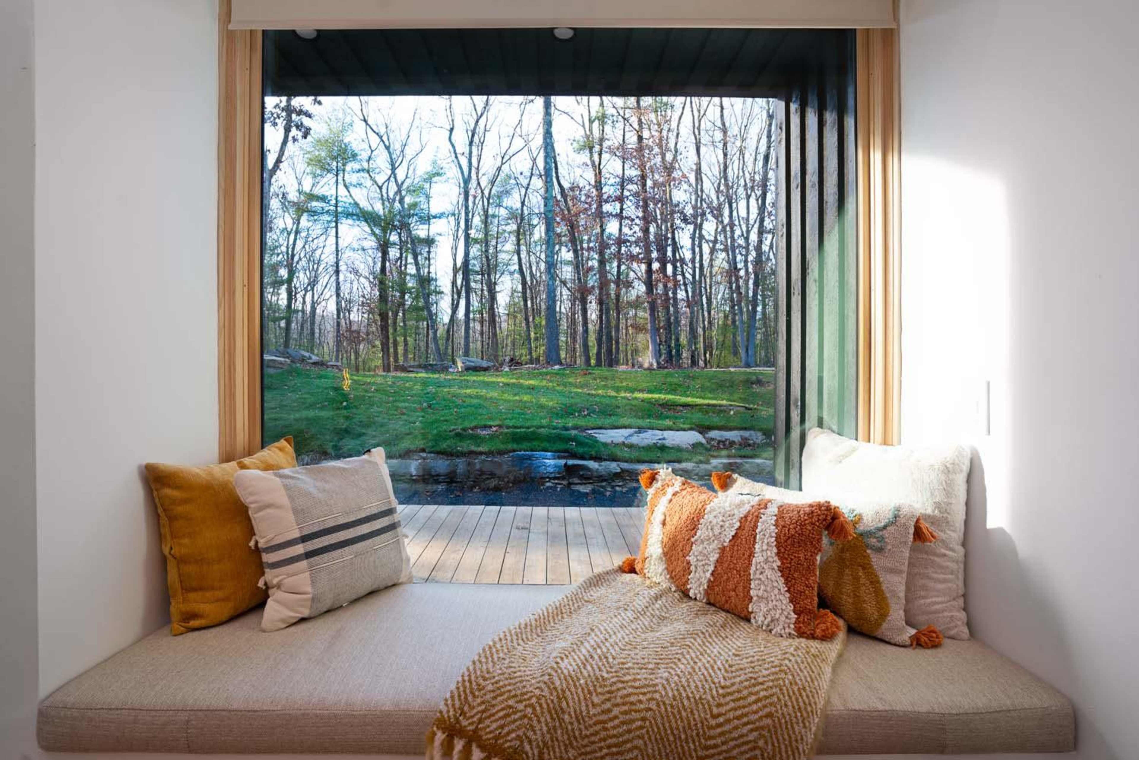 A cozy window seat with cushions overlooks a wooded landscape through large glass panes.