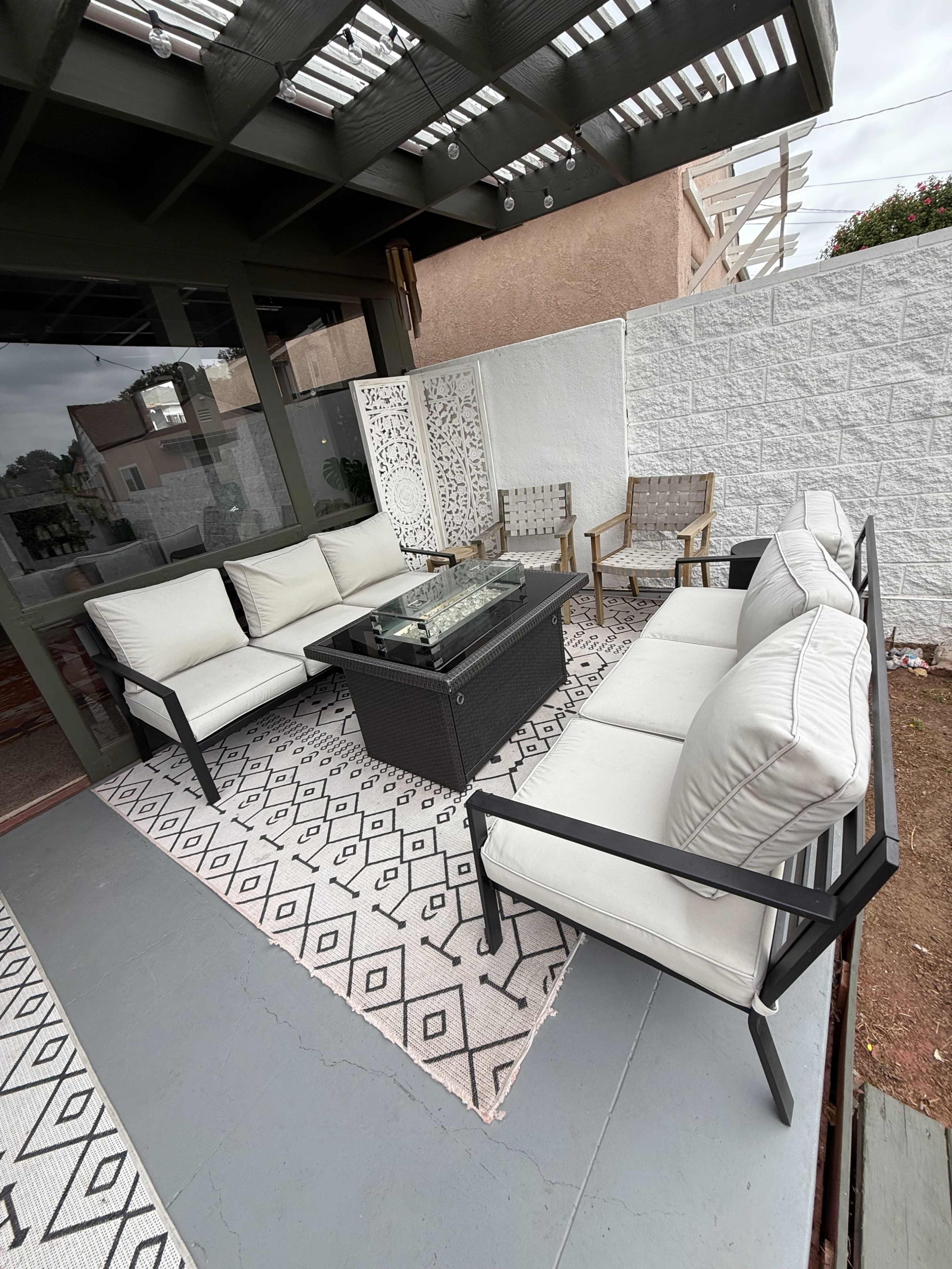 The image shows a patio area with a rectangular glass-top coffee table surrounded by a six-piece seating arrangement of white cushions and a patterned rug.