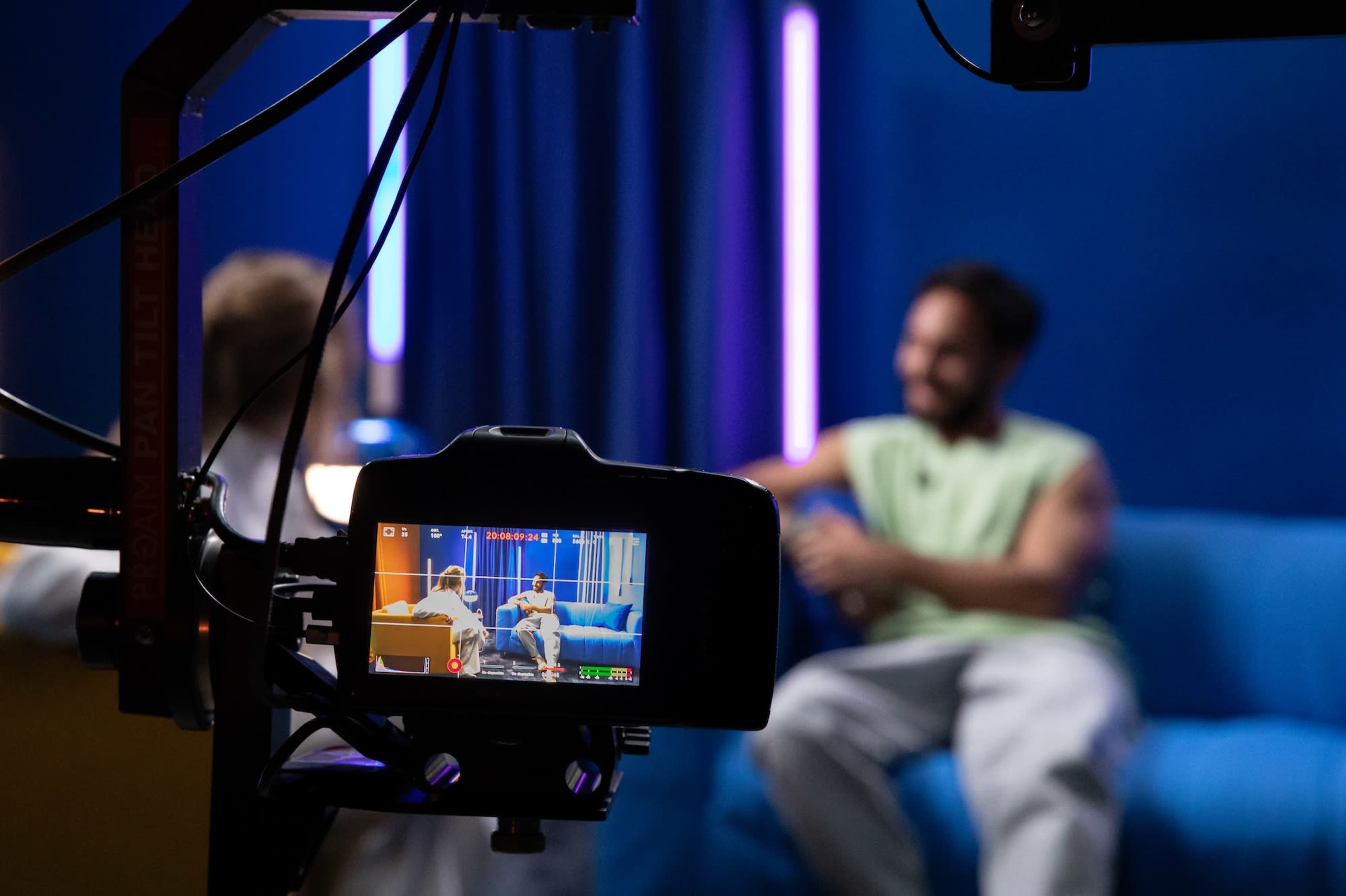 A video camera captures a conversation between two people seated on a blue couch, with colorful lights illuminating the background.