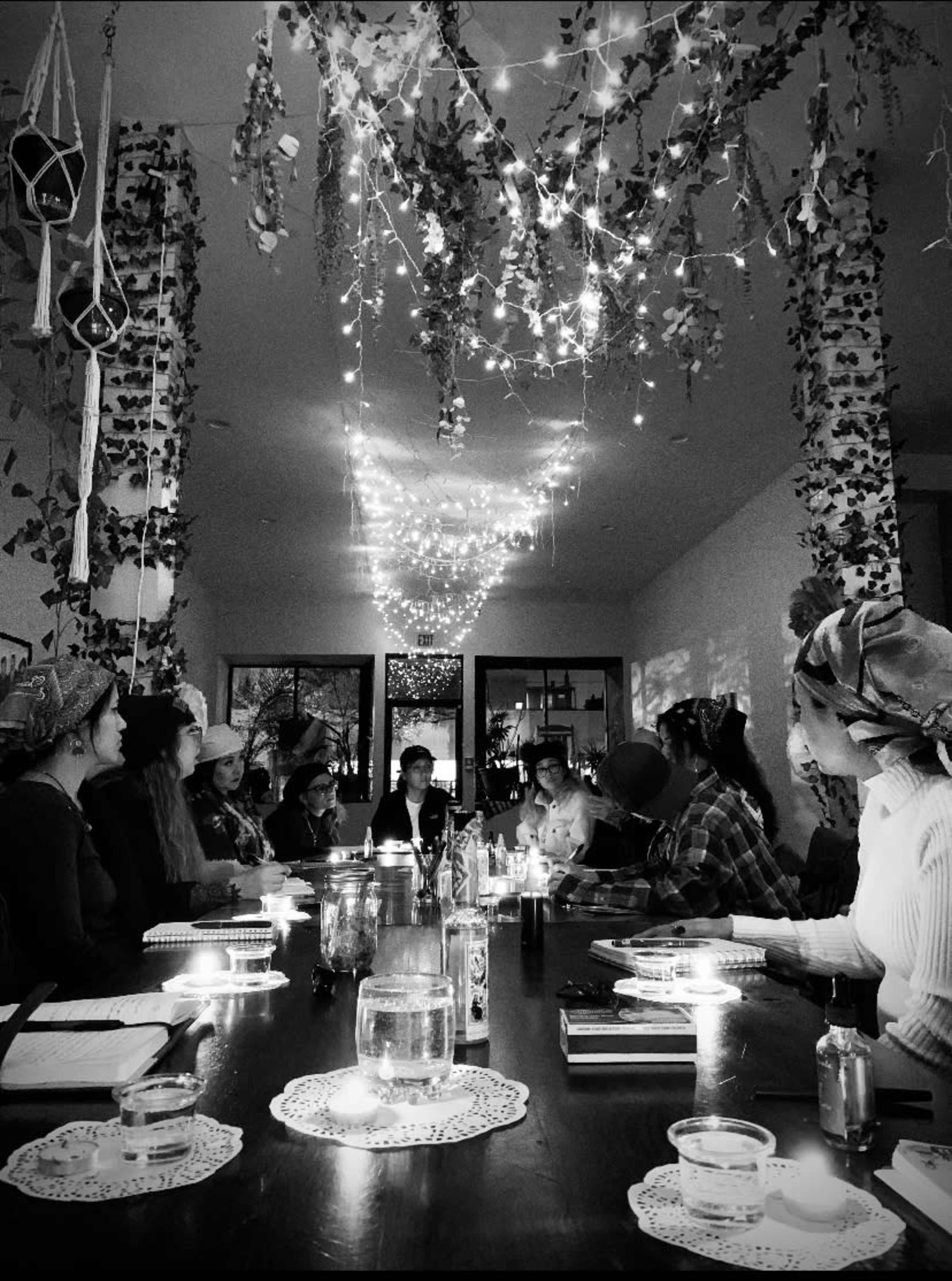 A group of people sits around a long table adorned with candles and foliage, while strings of lights hang above in a warmly lit room.