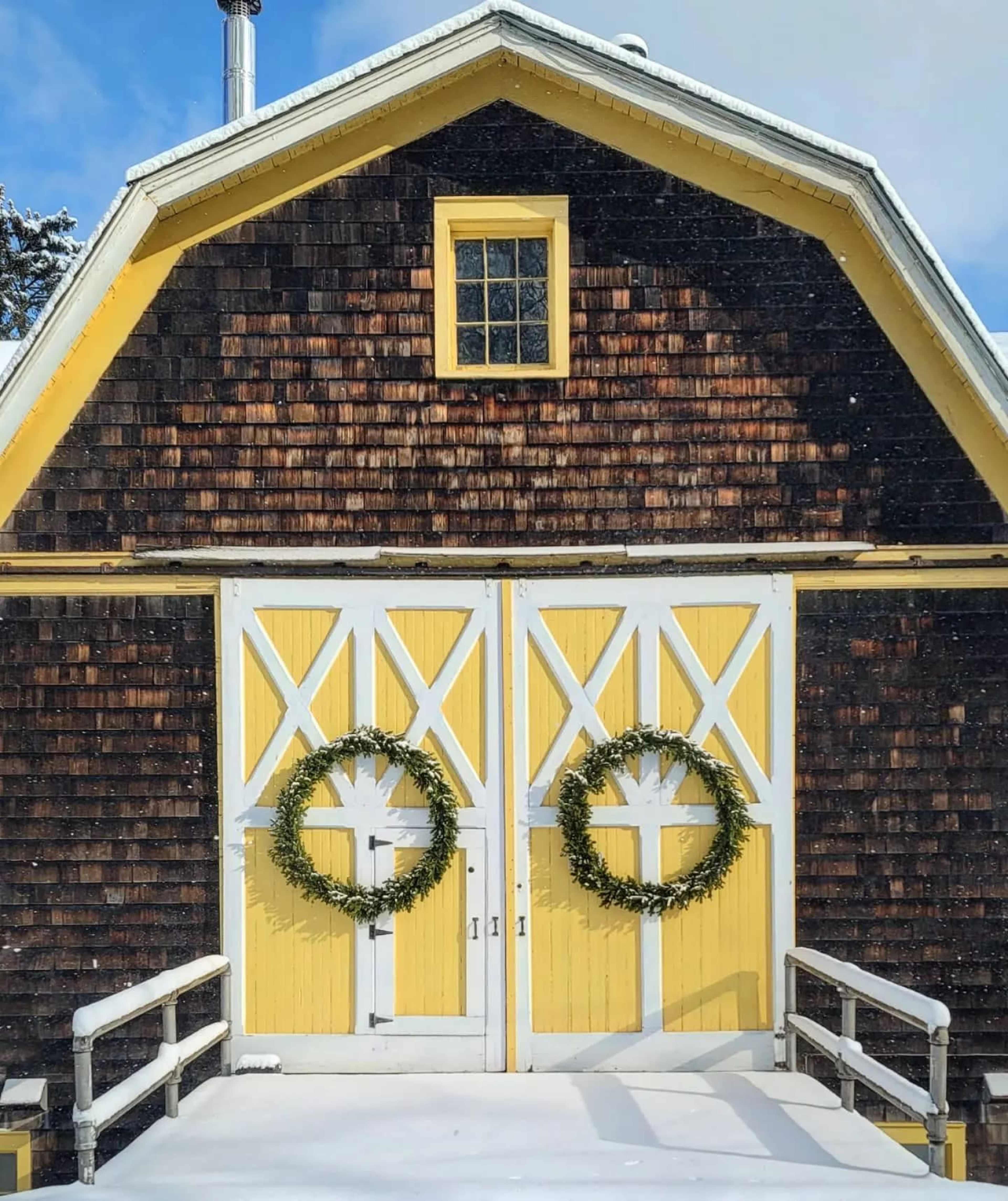A rustic barn with a yellow facade and two circular wreaths on the front doors, set against a snowy landscape.
