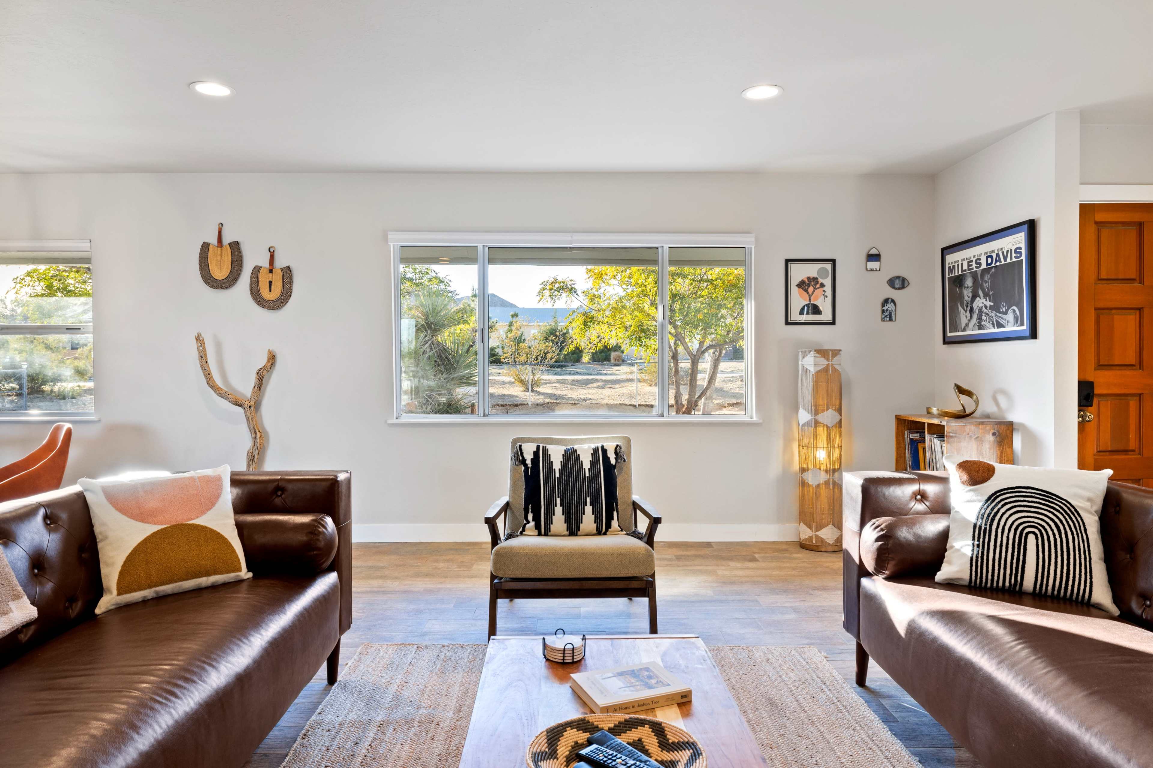 The image shows a modern living room with leather sofas, a patterned armchair, a wooden coffee table, and a large window overlooking a scenic view.