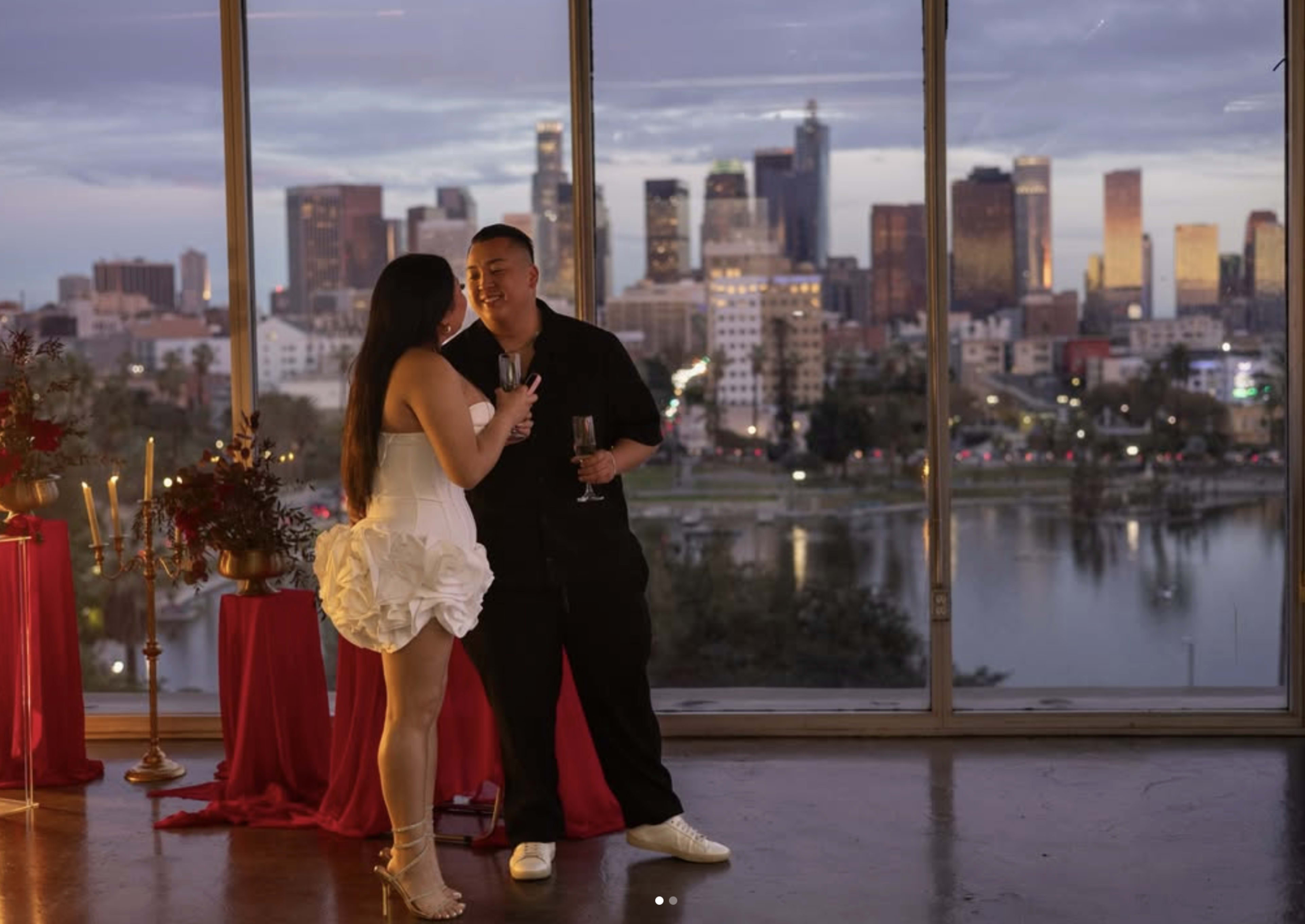 A couple stands together, holding glasses, against a backdrop of a city skyline at sunset.