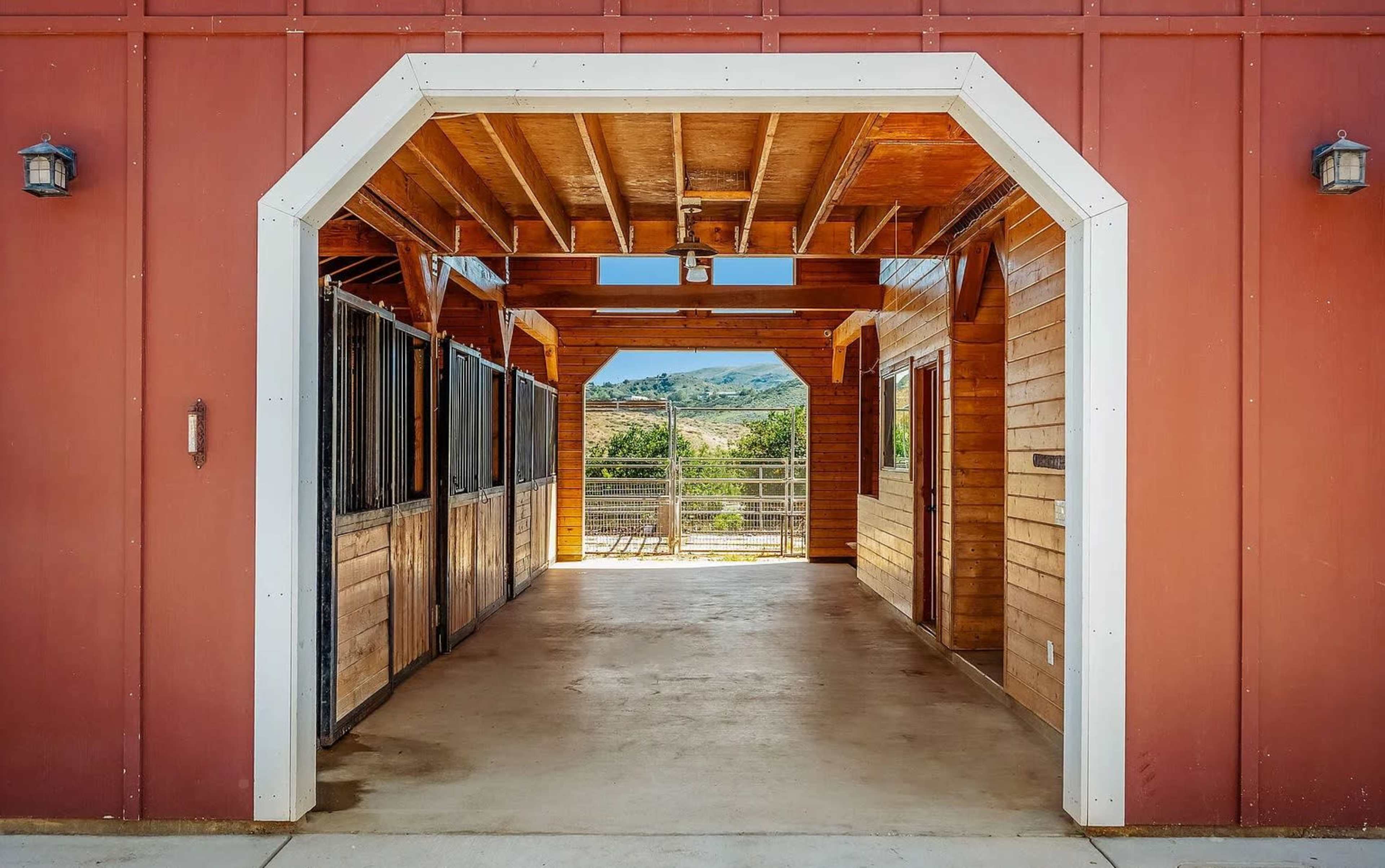 The image shows the interior of a red barn structure with wooden stalls on either side and an open entrance leading to a view of hills in the background.