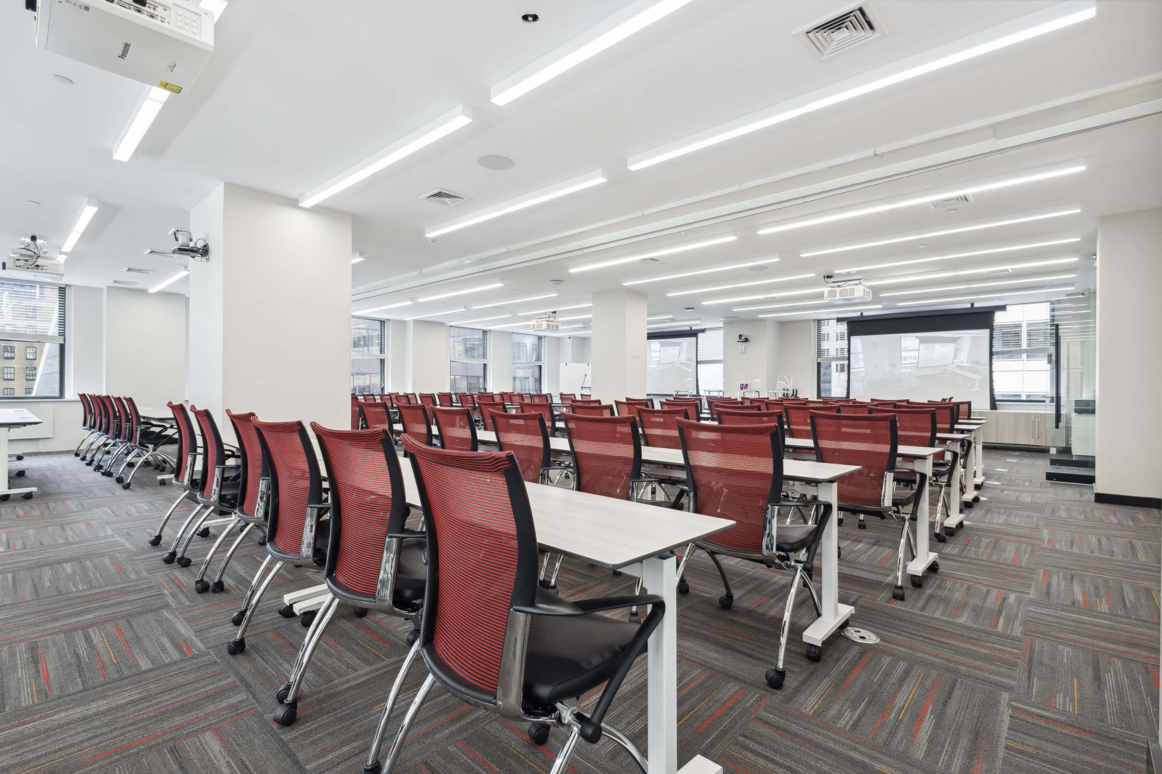 A modern classroom is arranged with rows of red and gray chairs and tables, featuring multiple projectors and large windows for natural light.