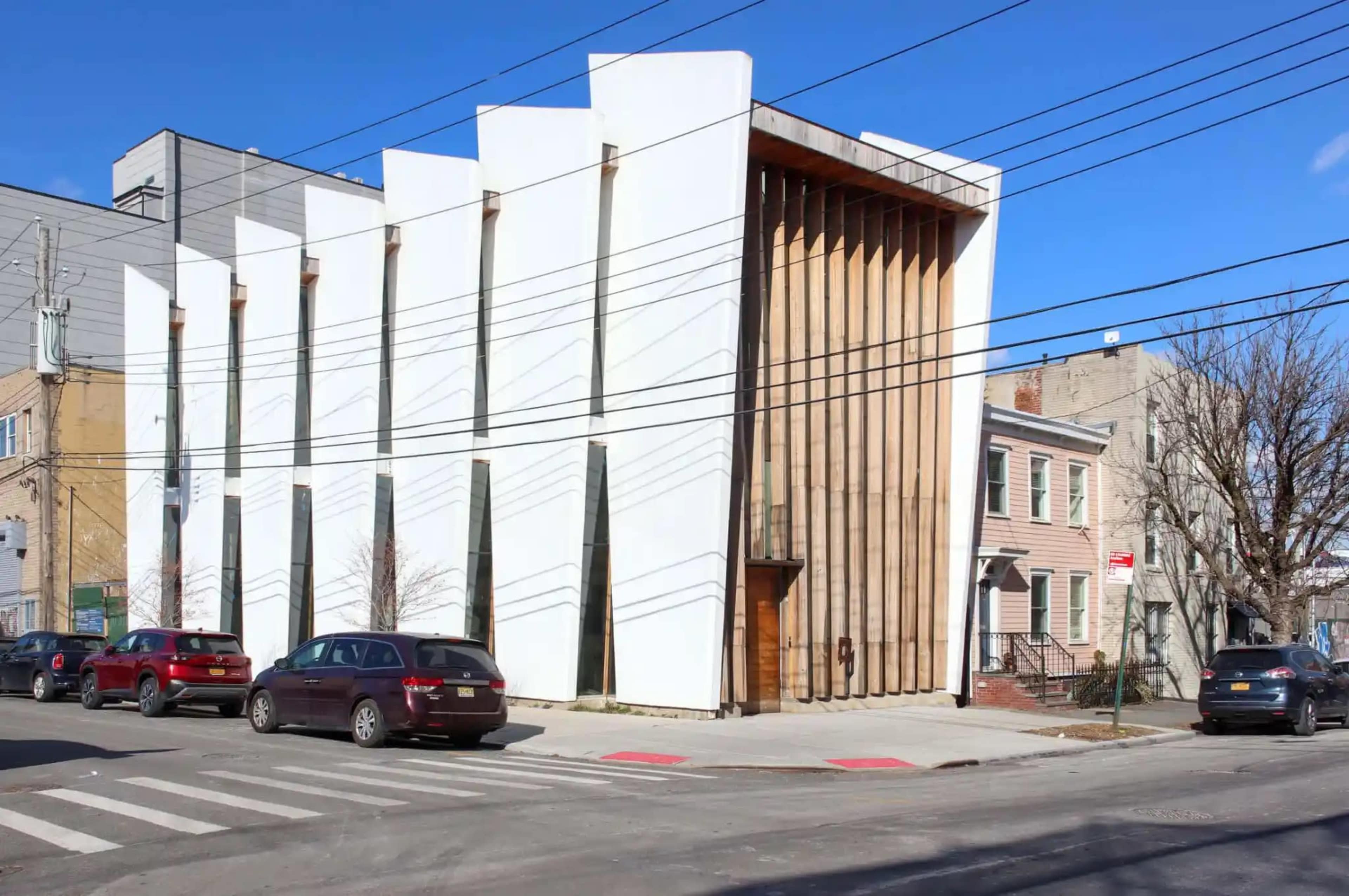 The image shows a modern building with a striking white facade and wooden accents, situated at a street corner next to traditional houses and parked cars.