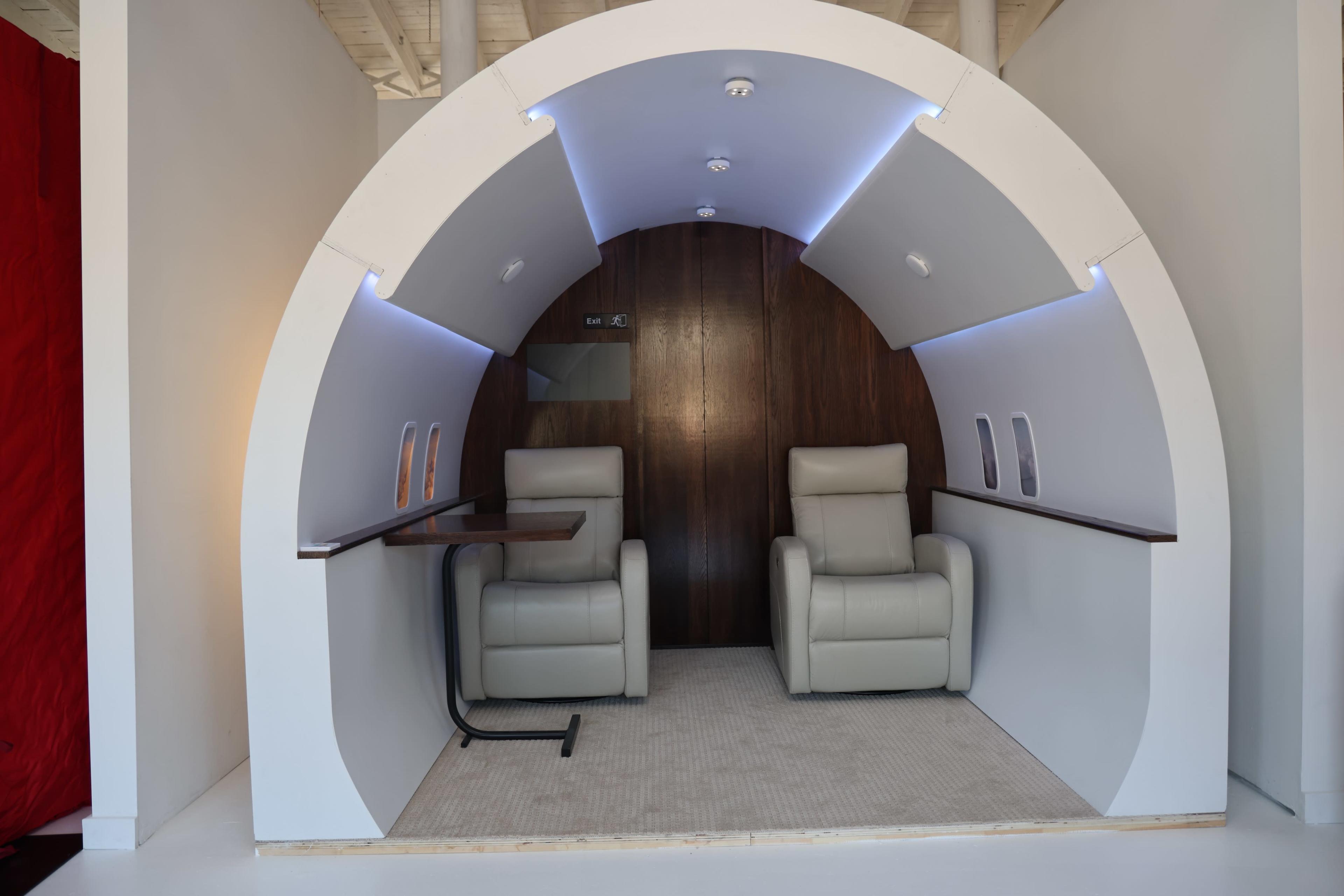 A minimalist lounge area resembling an aircraft cabin, featuring two leather armchairs and a small table under a curved ceiling with recessed lighting.