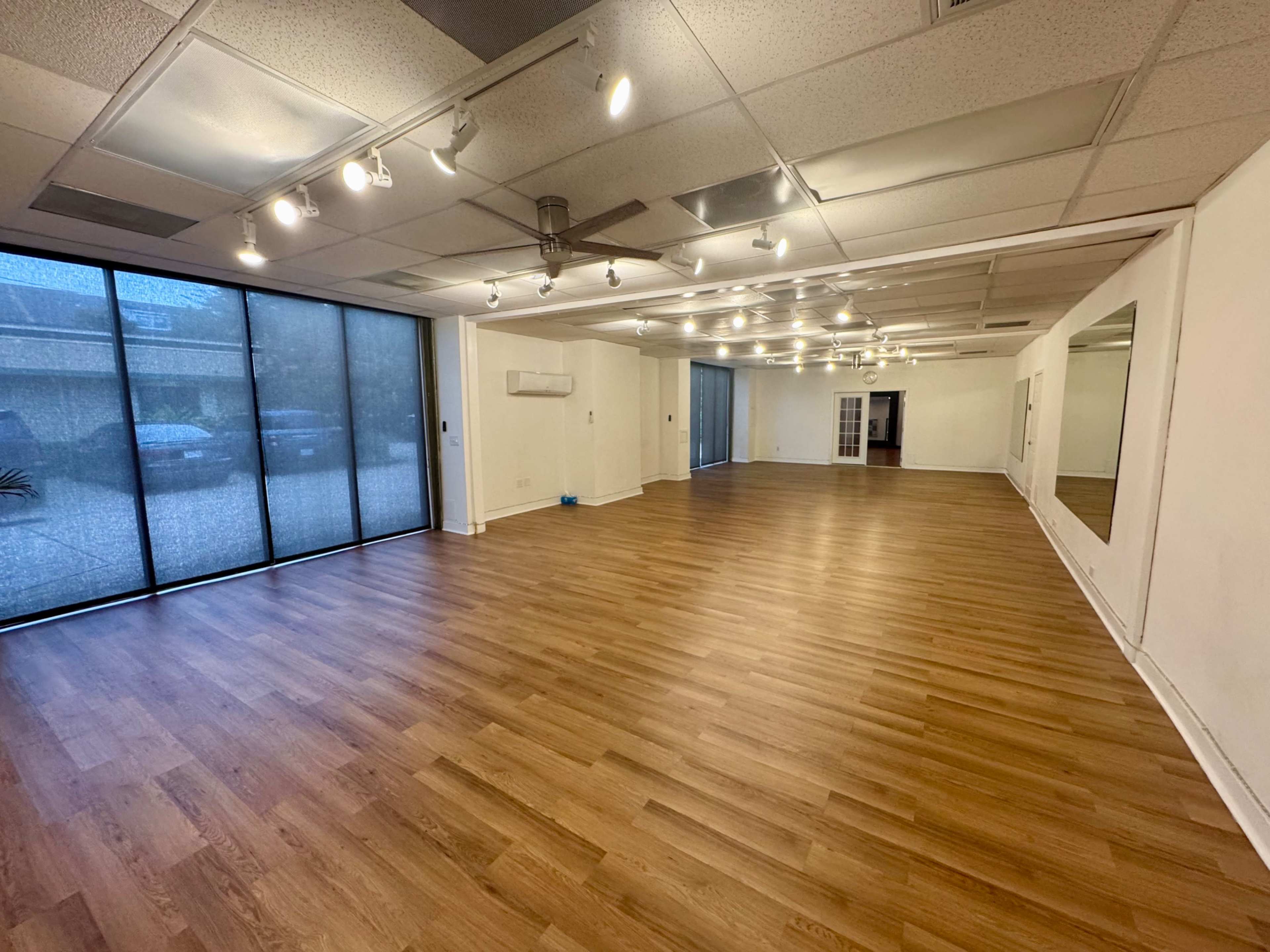 The image shows a spacious, empty room with wooden flooring and large windows allowing natural light to filter in.