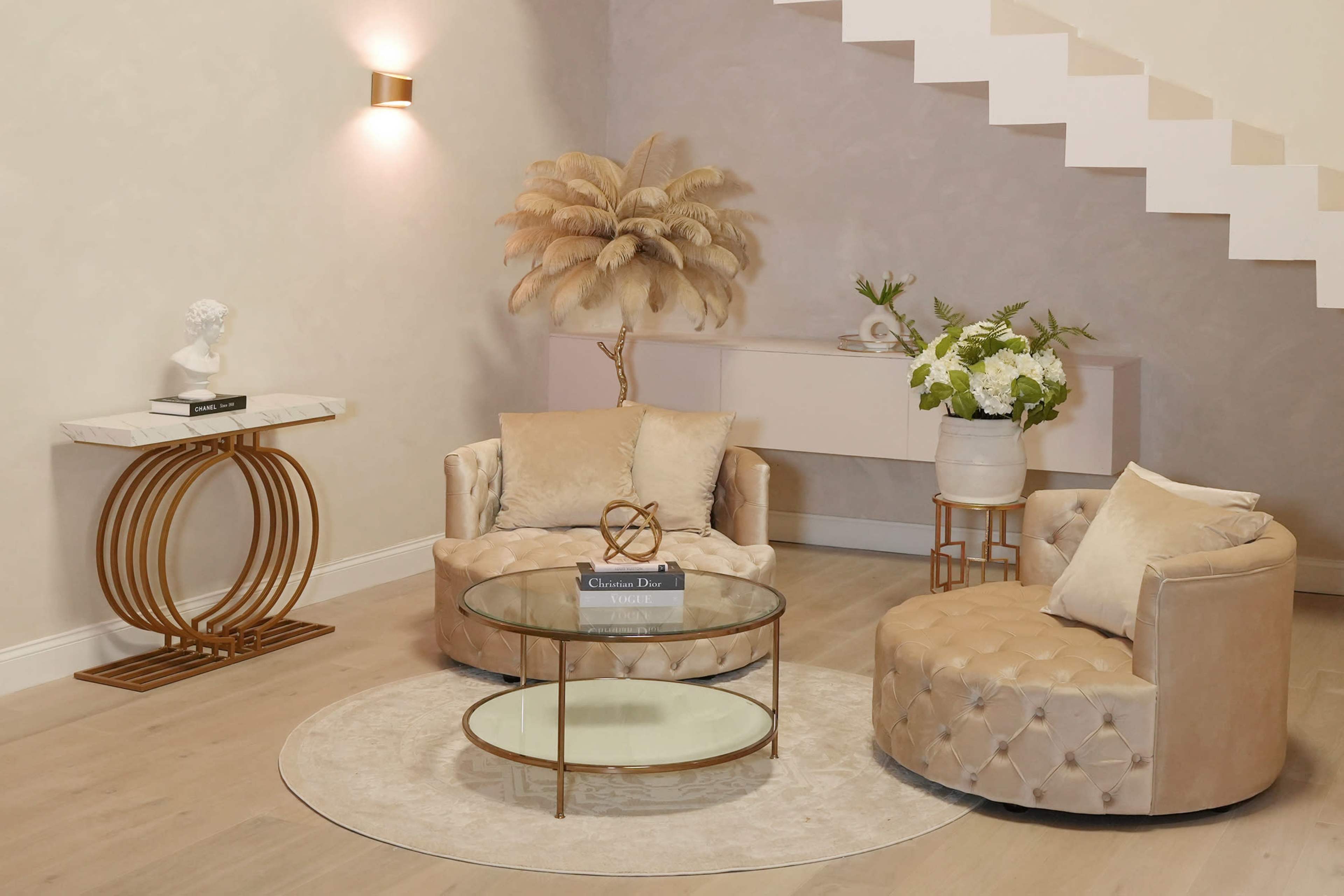 A modern living room setup featuring two upholstered chairs, a glass coffee table, and decorative elements like a statue and a potted plant against a light-colored wall.