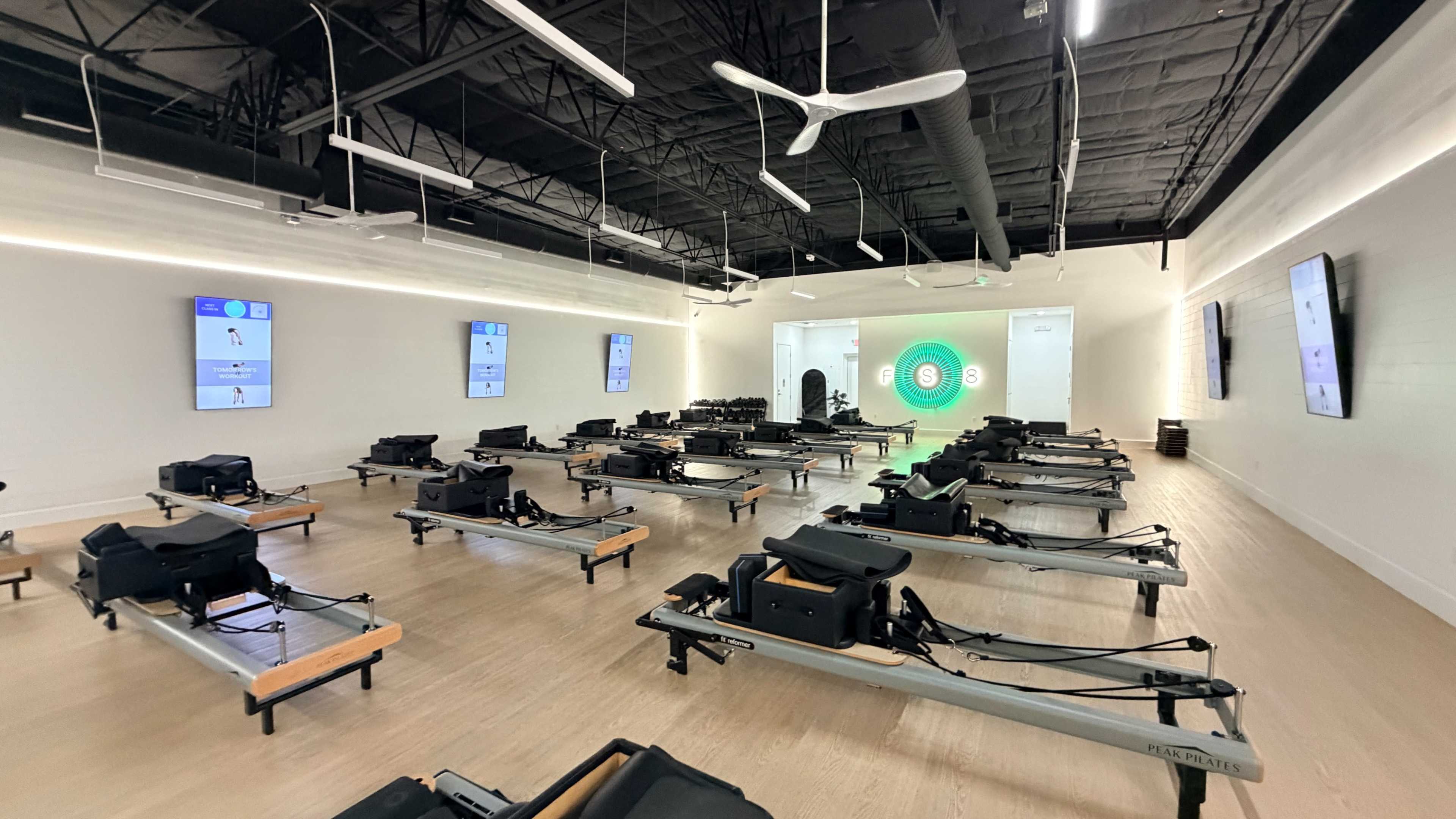 The image shows a spacious fitness studio equipped with multiple reformer Pilates machines arranged in rows, with large screens displaying workout information on the walls.