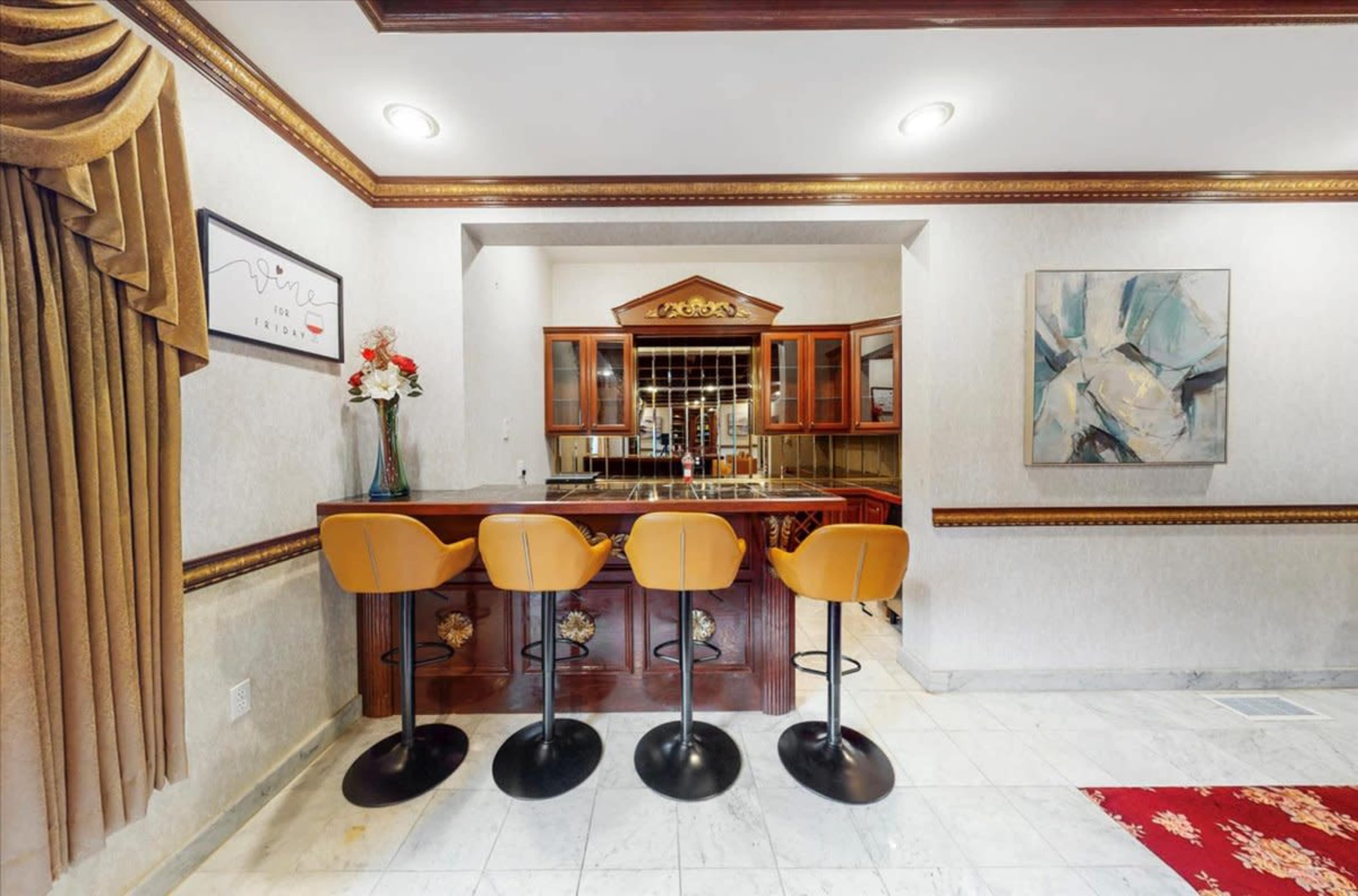 The image shows a home bar area with four black stools positioned in front of a wooden bar counter, surrounded by decorative walls and a large framed artwork.