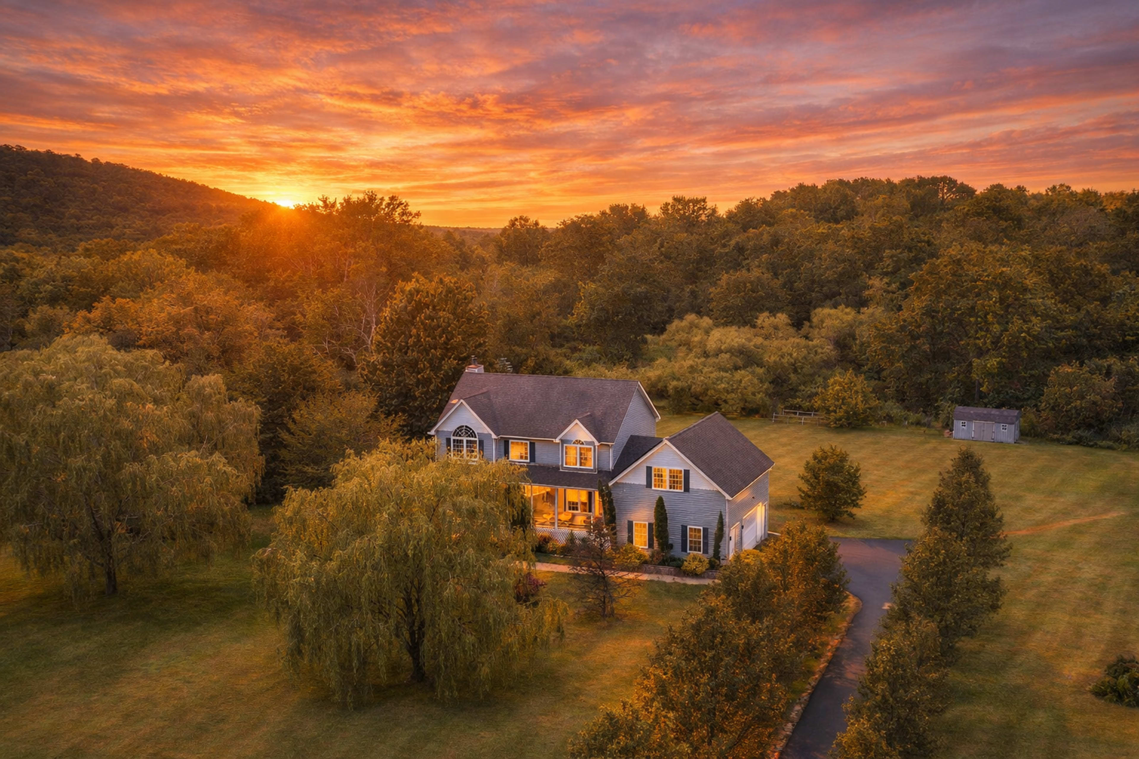 A large, two-story house with a front porch is situated in a green landscape at sunset, surrounded by trees and fields.