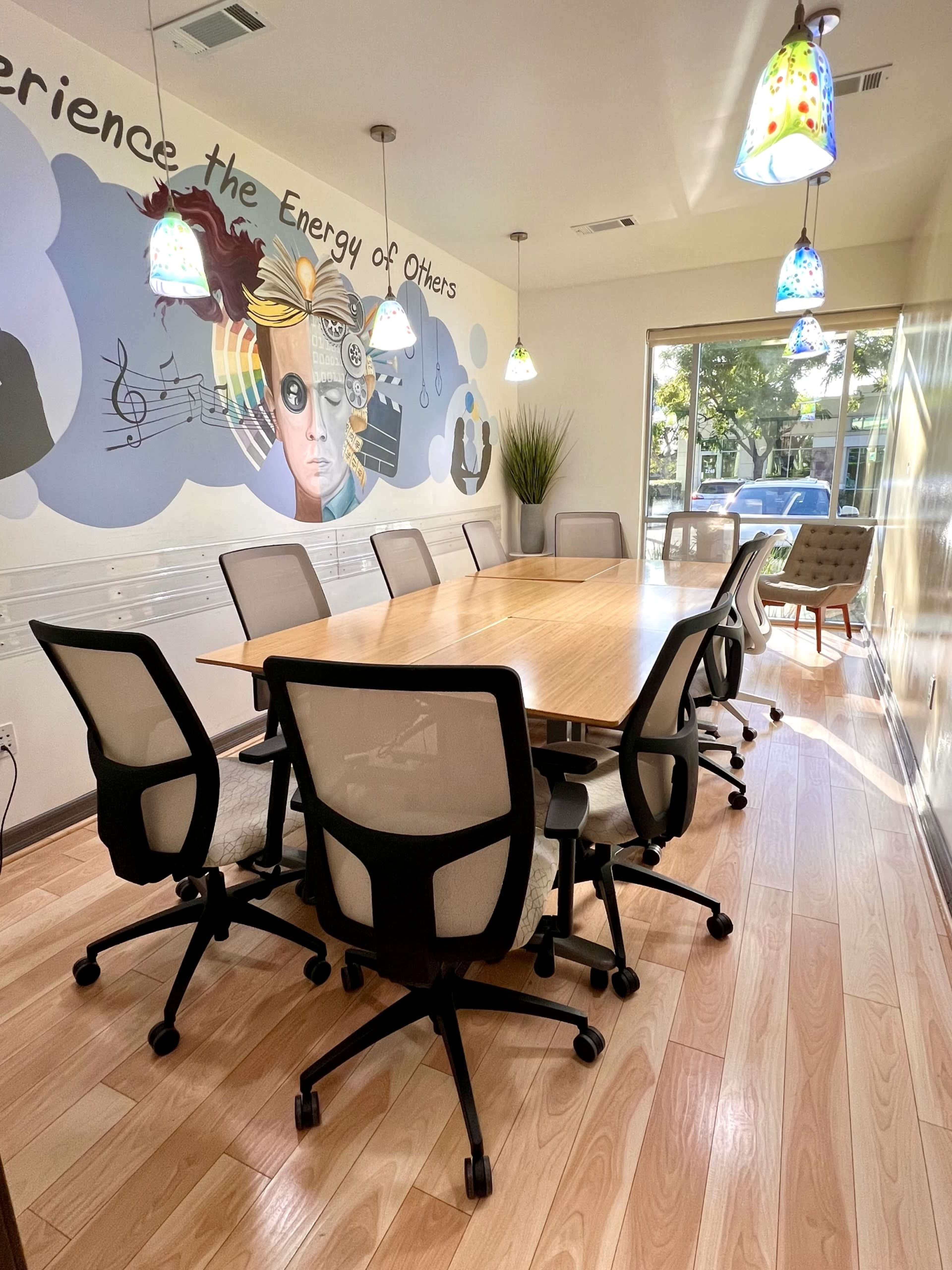 A brightly lit conference room features a long wooden table surrounded by several office chairs and a mural on the wall.