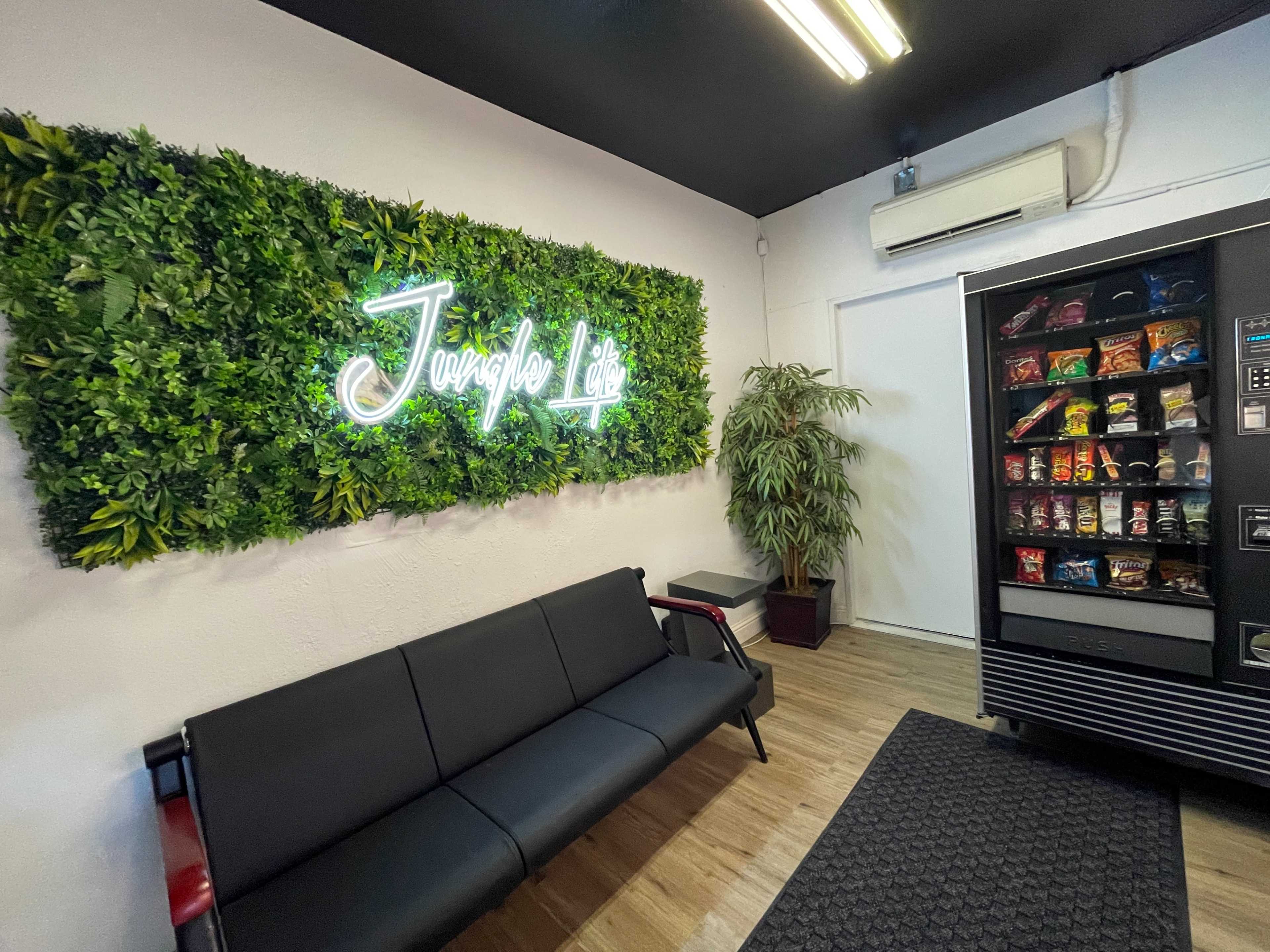 The image shows a seating area with a black couch, a green plant wall featuring a neon sign that reads "Jungle Life," and a vending machine stocked with snacks and drinks.