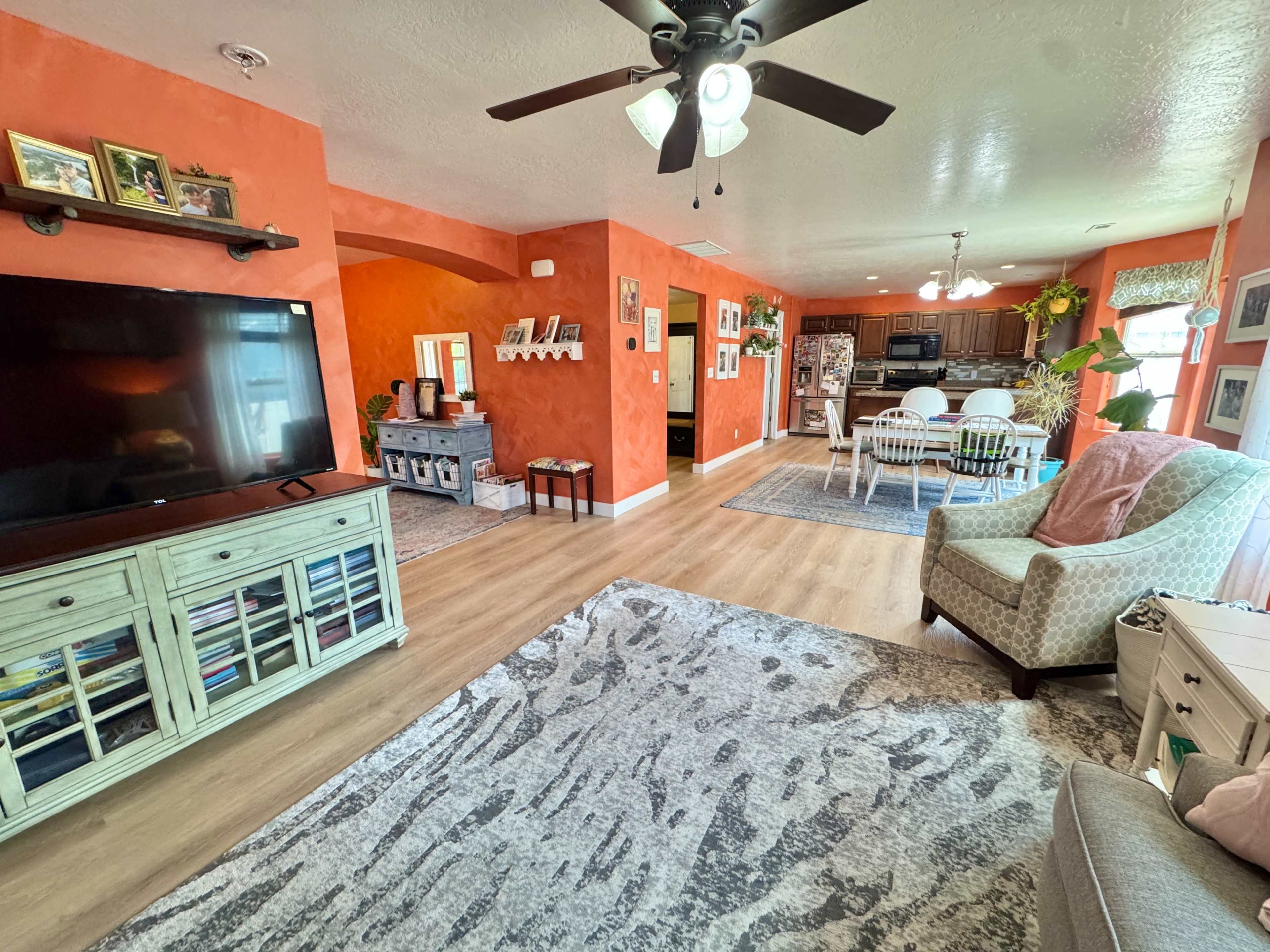 The image shows a living room with orange walls, a television stand, a patterned rug, and an open view into a kitchen and dining area.