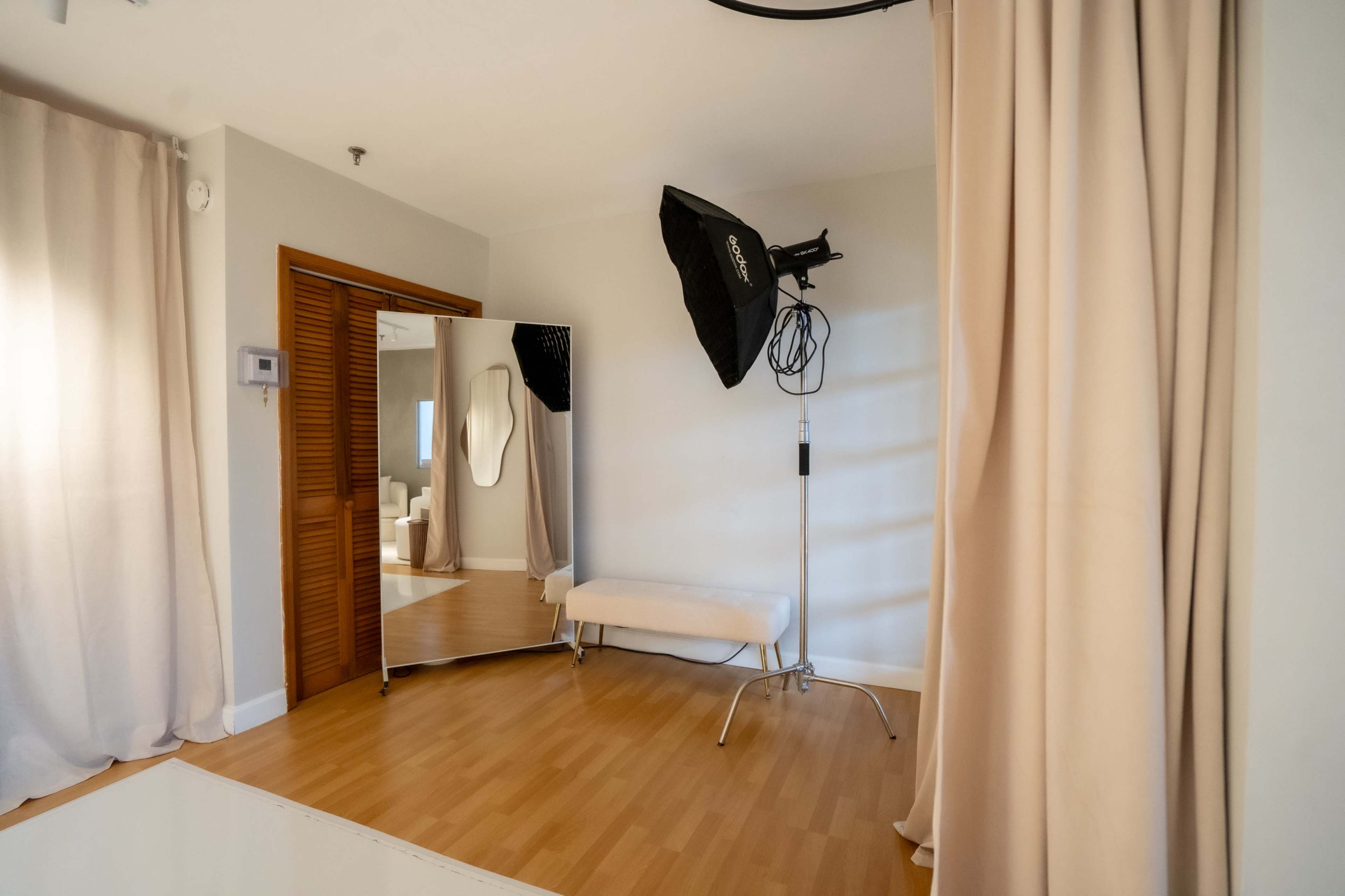 The image shows a minimalist photography studio featuring a large mirror, a softbox light on a stand, a bench, and beige curtains.
