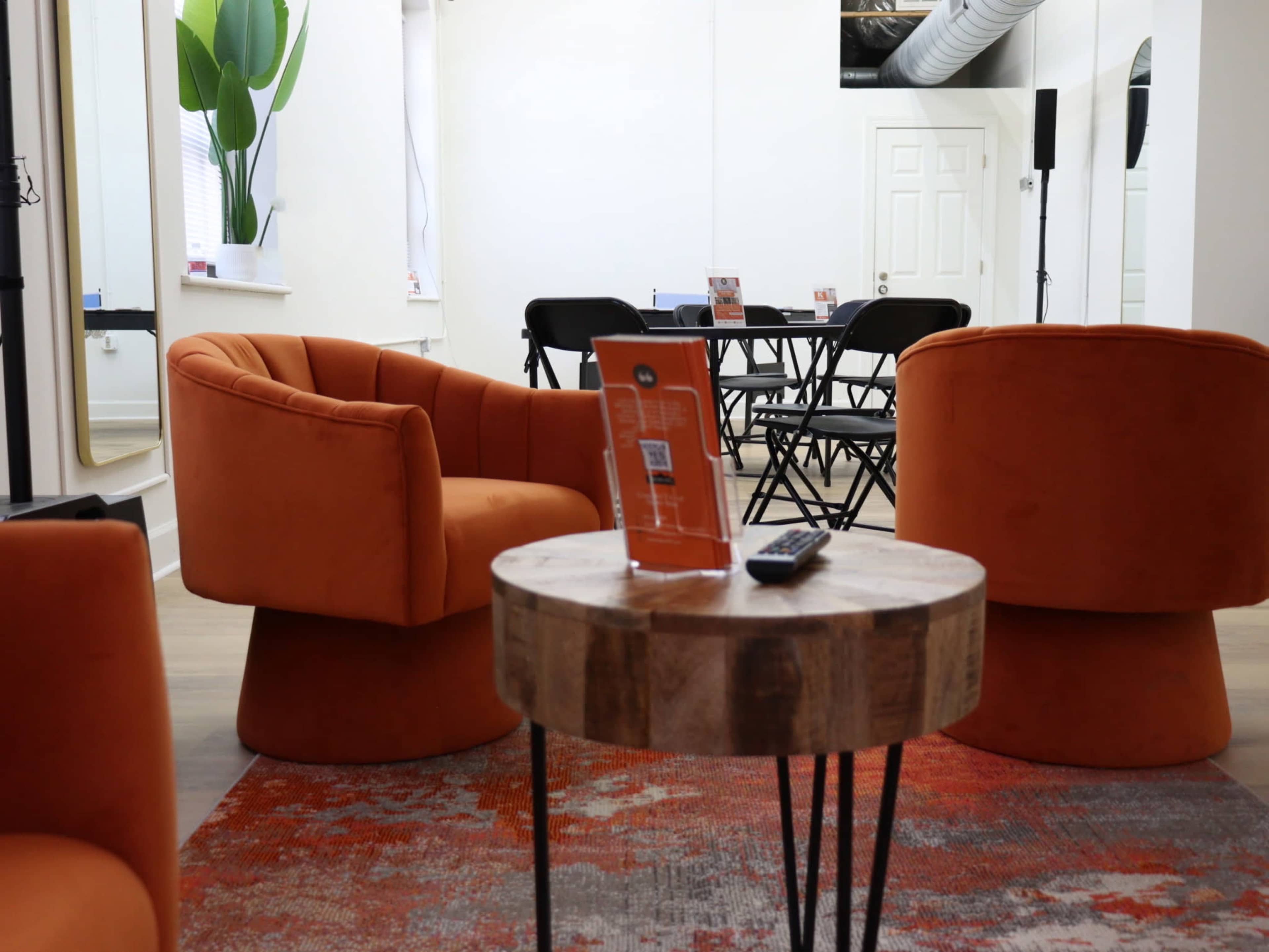 The image shows two orange upholstered chairs facing a small wooden table on a patterned rug in a modern, brightly lit room with a folding table and chairs in the background.