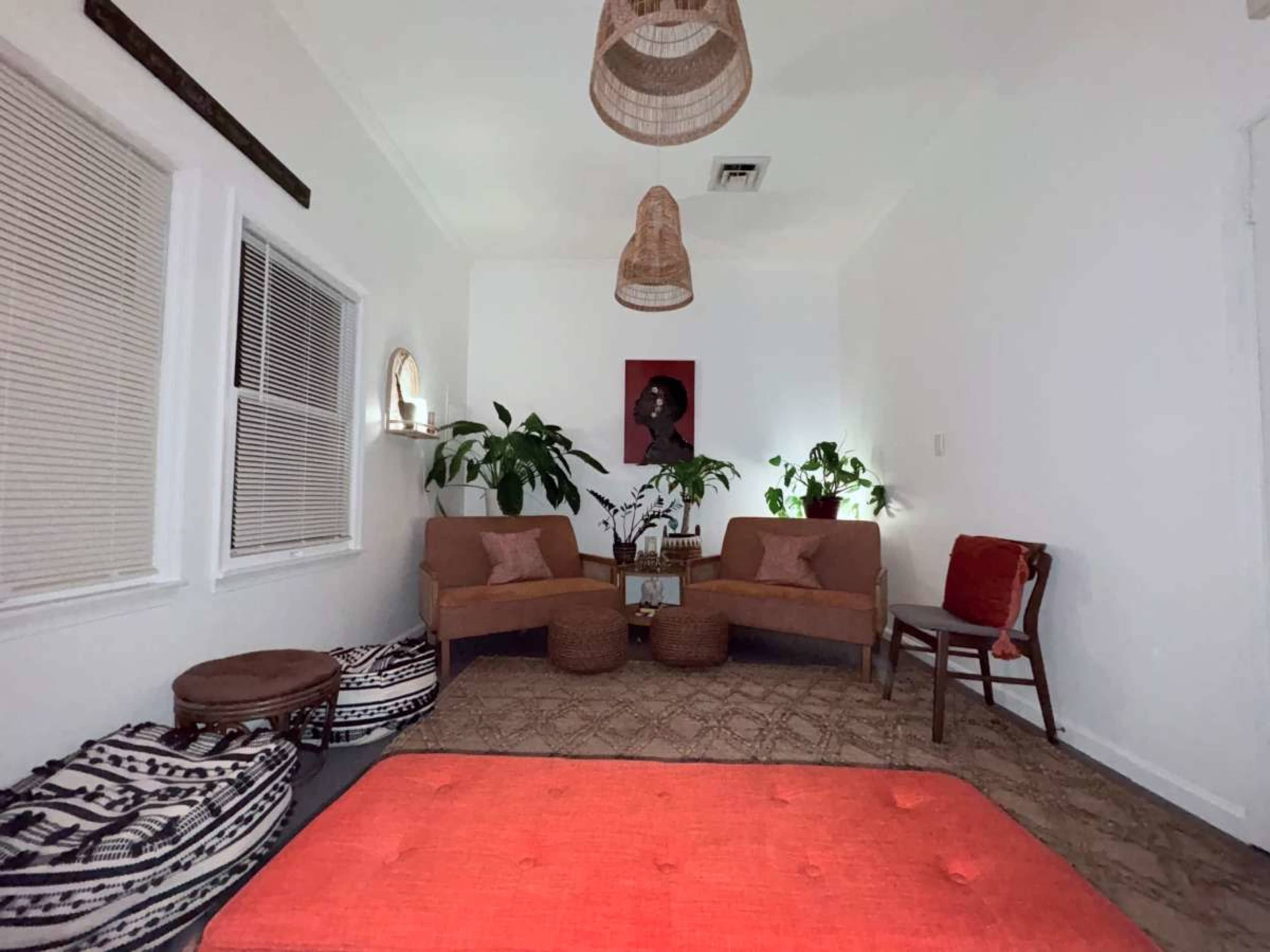 The image depicts a cozy, minimalist living room featuring two upholstered chairs, a small table with plants, and a large red rug on a patterned floor.