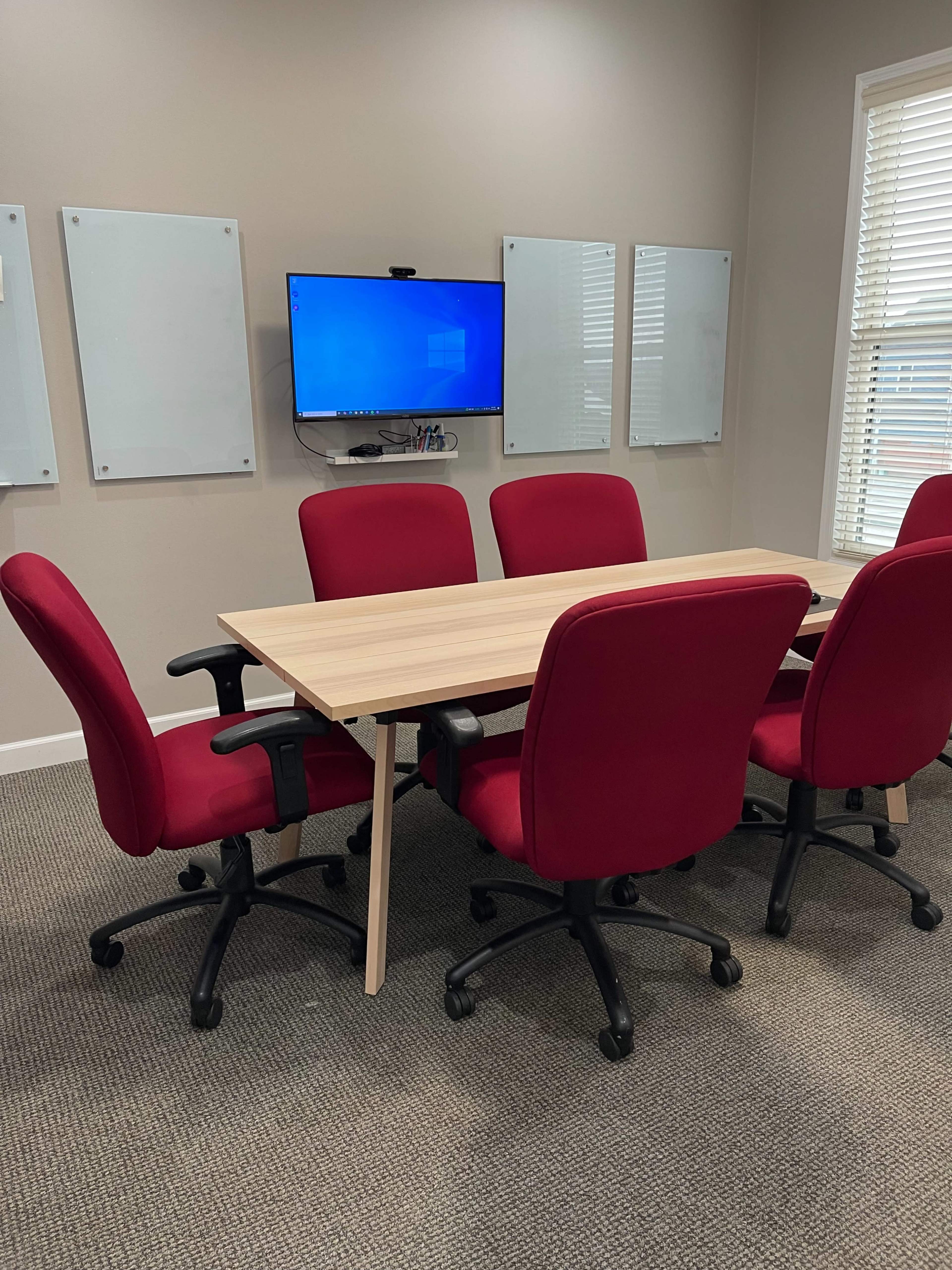 A conference room features a rectangular wooden table surrounded by four red office chairs, with a television mounted on the wall and whiteboards positioned nearby.
