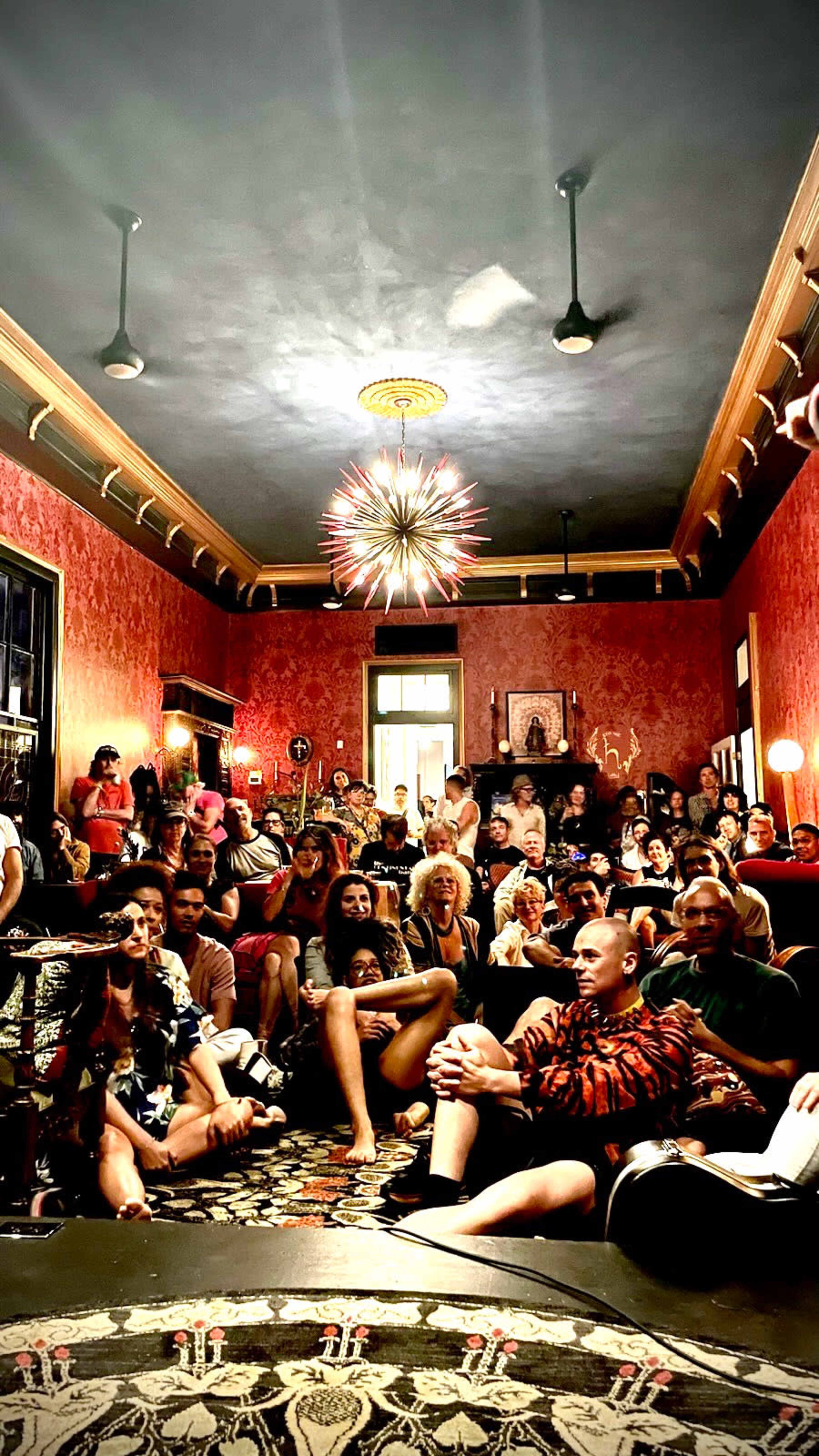 A large group of people gathers in a richly decorated room with red walls and ornate lighting fixtures.