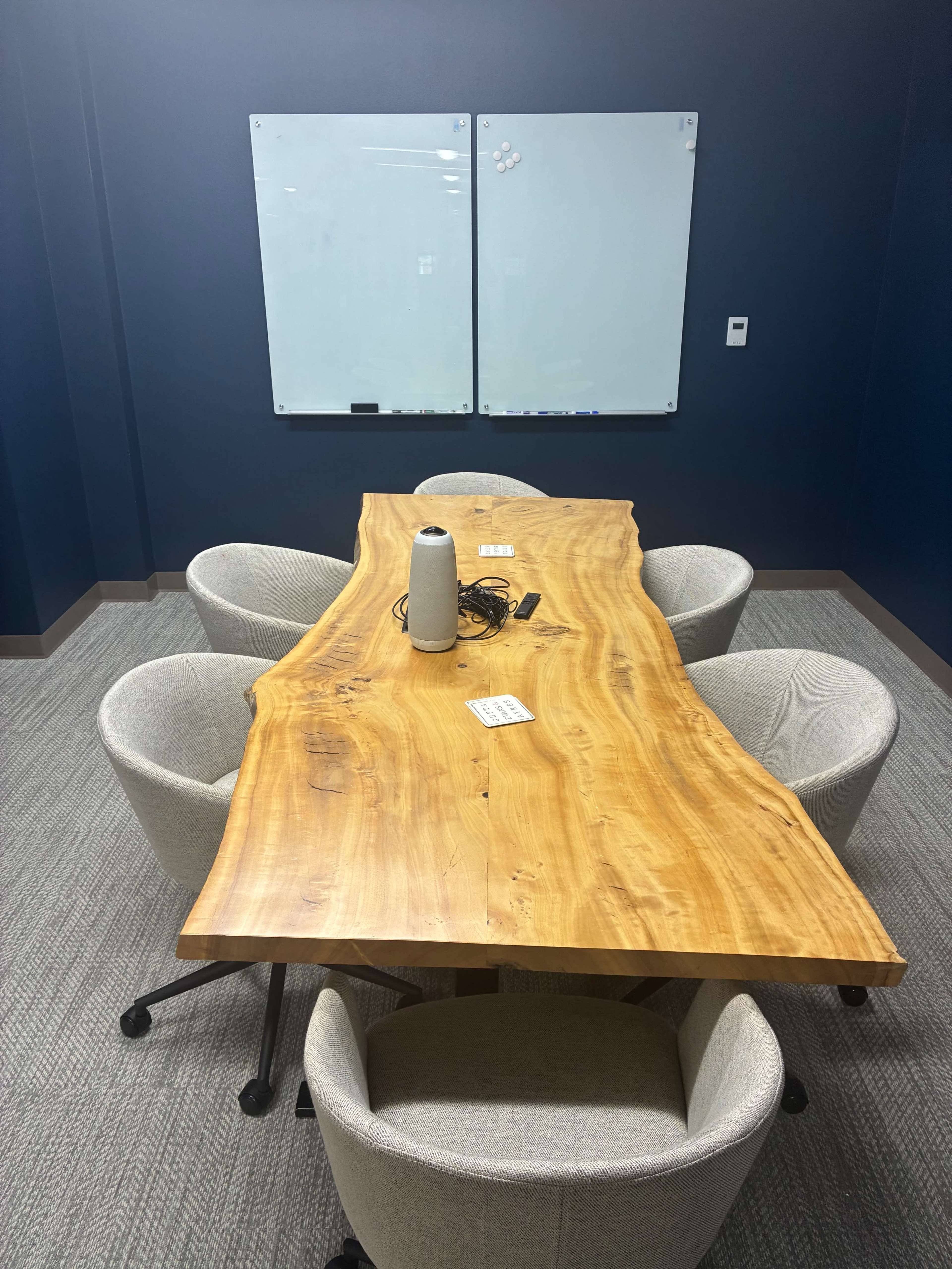 A modern meeting room features a large wooden table surrounded by four gray chairs, with two whiteboards mounted on the wall.