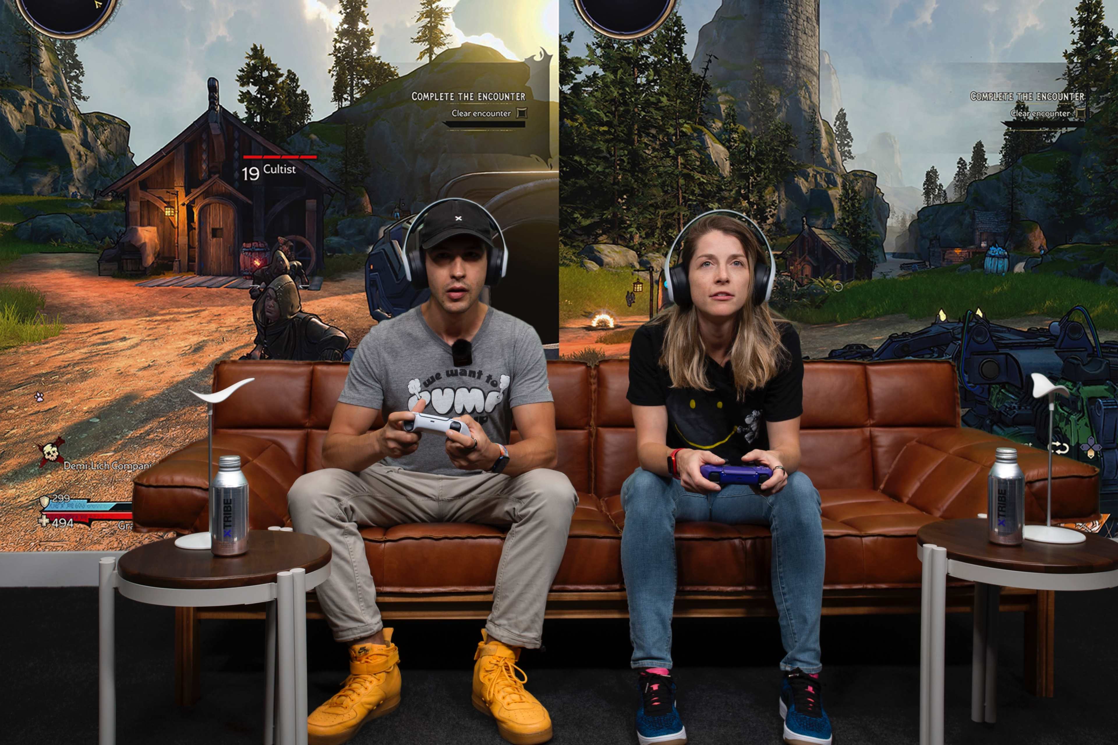 Two individuals are seated on a brown sofa, wearing headsets and playing video games on connected consoles in front of a large display showing game graphics.