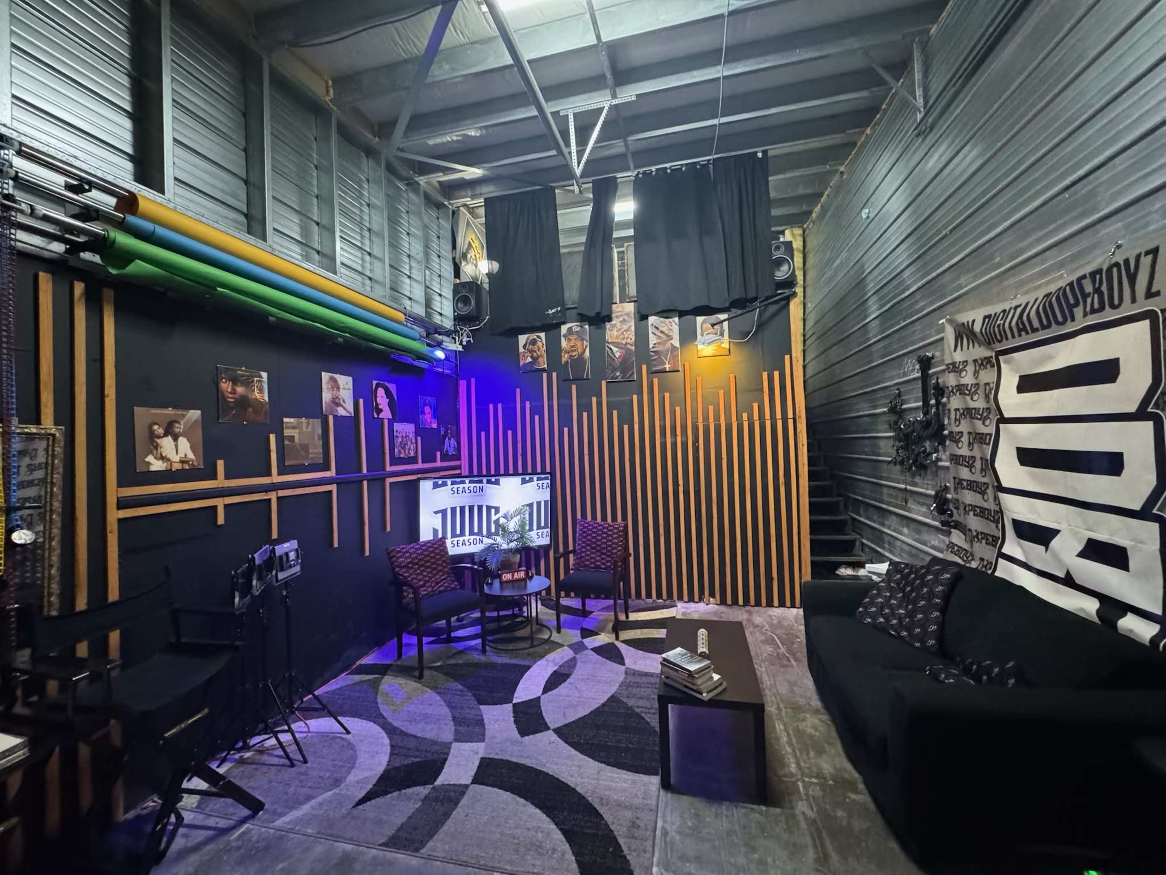 The image shows a cozy recording studio or lounge area featuring dark walls, a seating arrangement with two chairs and a couch, ambient lighting, and various framed pictures on the walls.