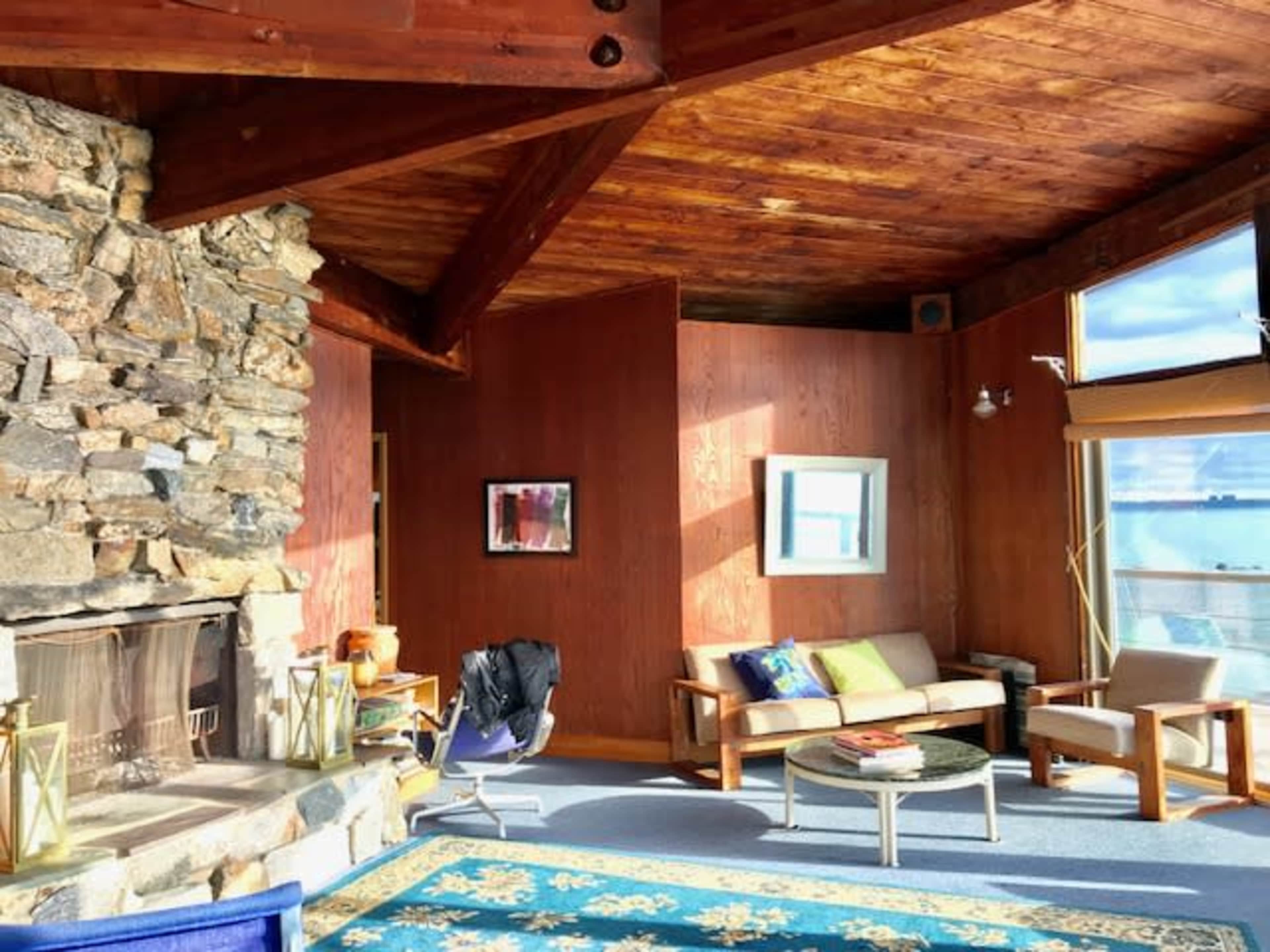 A wooden interior room with a stone fireplace, large windows, and a mix of seating arrangements, all situated near a body of water.