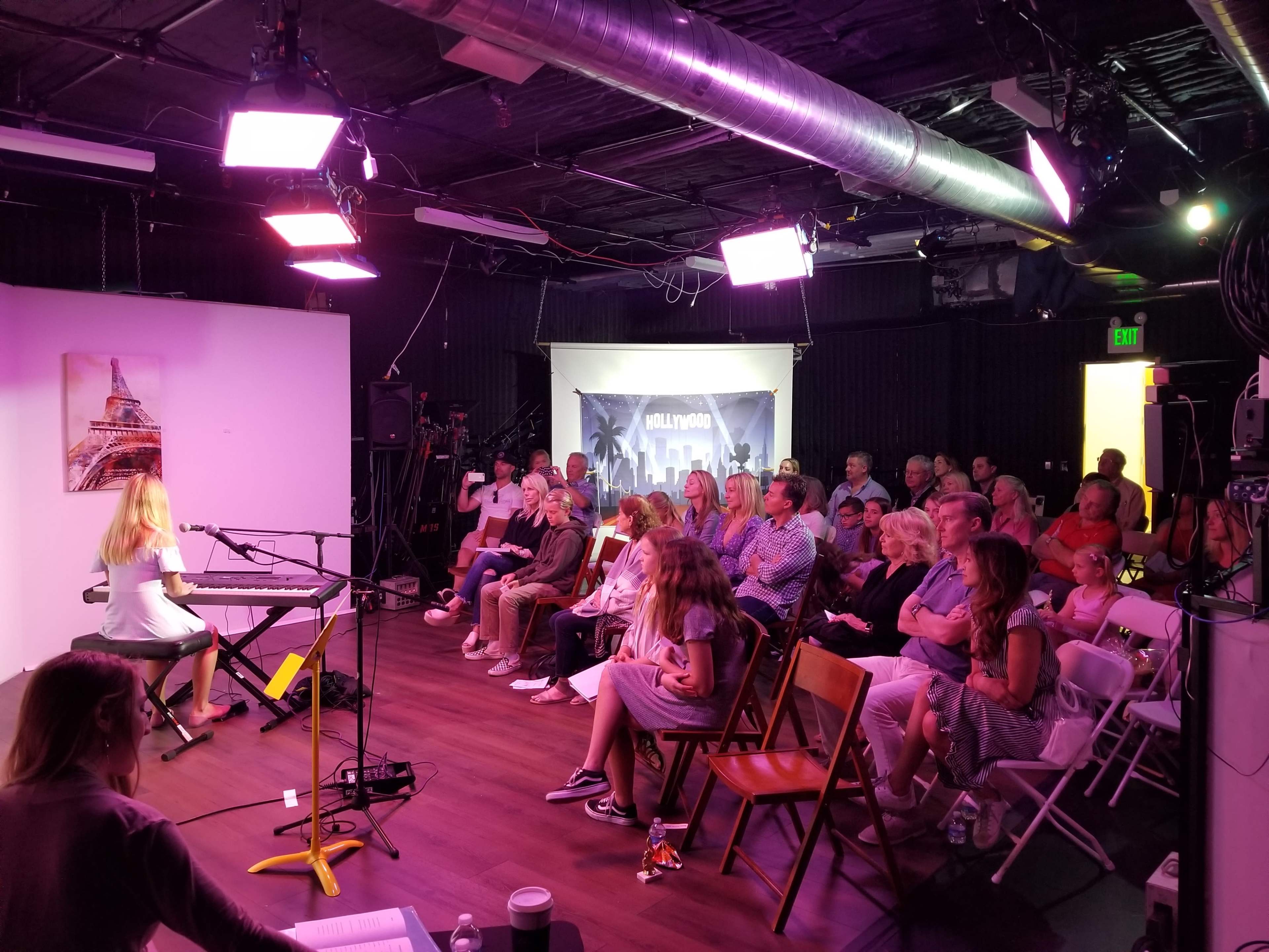 A performer plays a keyboard while an audience of seated spectators listens intently in a studio setting.