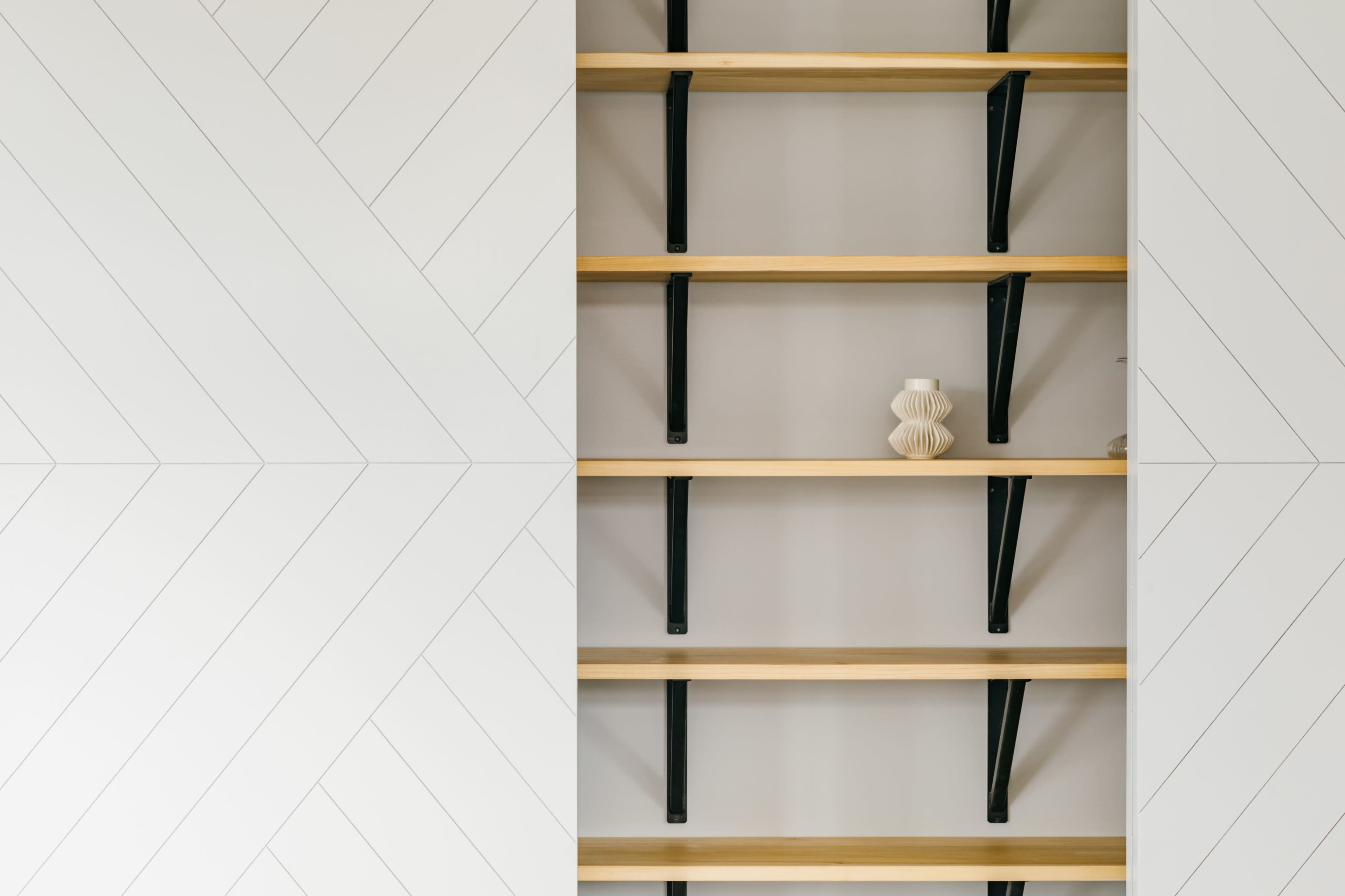 A row of wooden shelves mounted on a wall with a geometric design, featuring a single decorative vase on the center shelf.