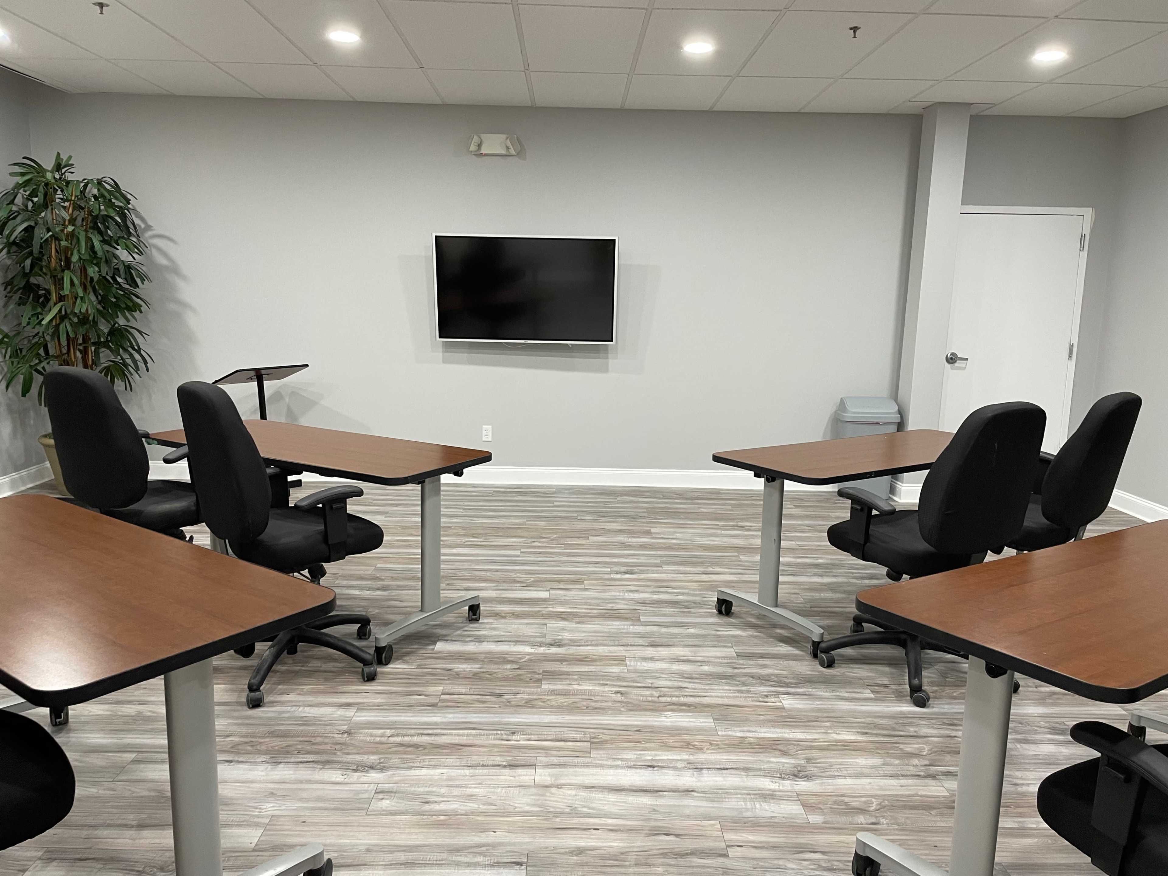 A meeting room features four tables with chairs arranged around them, facing a wall-mounted television, and includes a potted plant in the corner.
