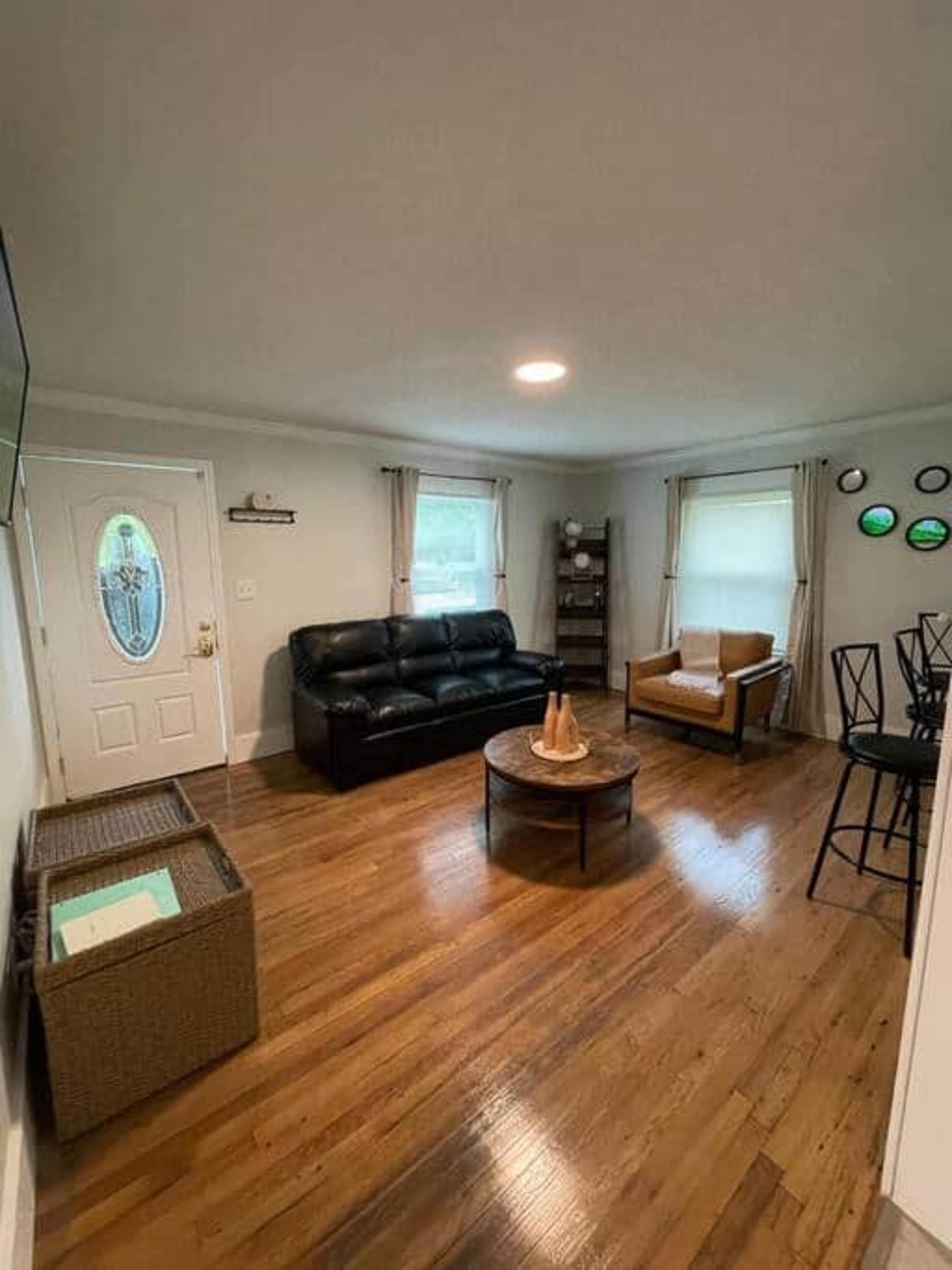 The room features hardwood floors, a black leather sofa, a brown armchair, a round coffee table, and a decorative shelf with circular wall decor.
